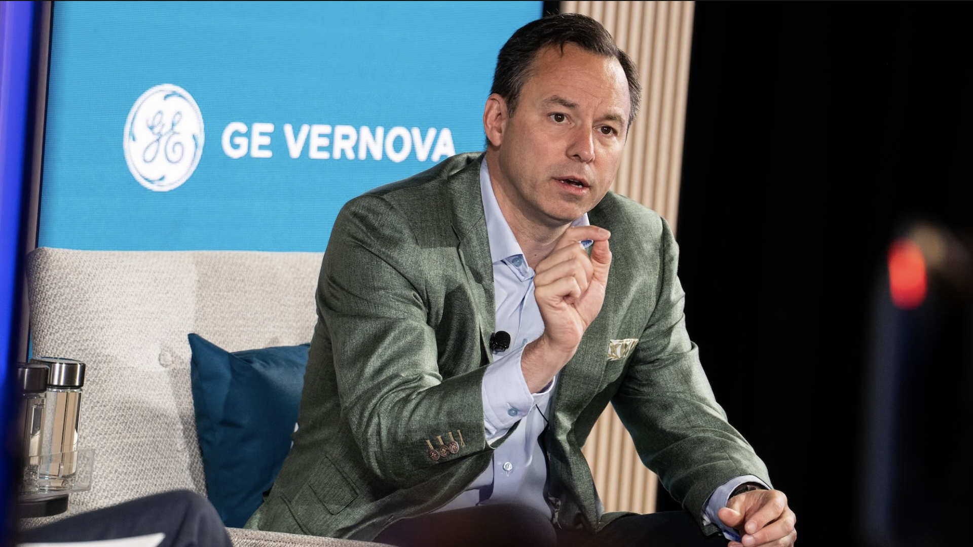 A man in a green blazer and light blue shirt speaking, gesturing with his hand, seated on a beige chair with blue pillow, blue screen behind him shows GE Vernova logo.