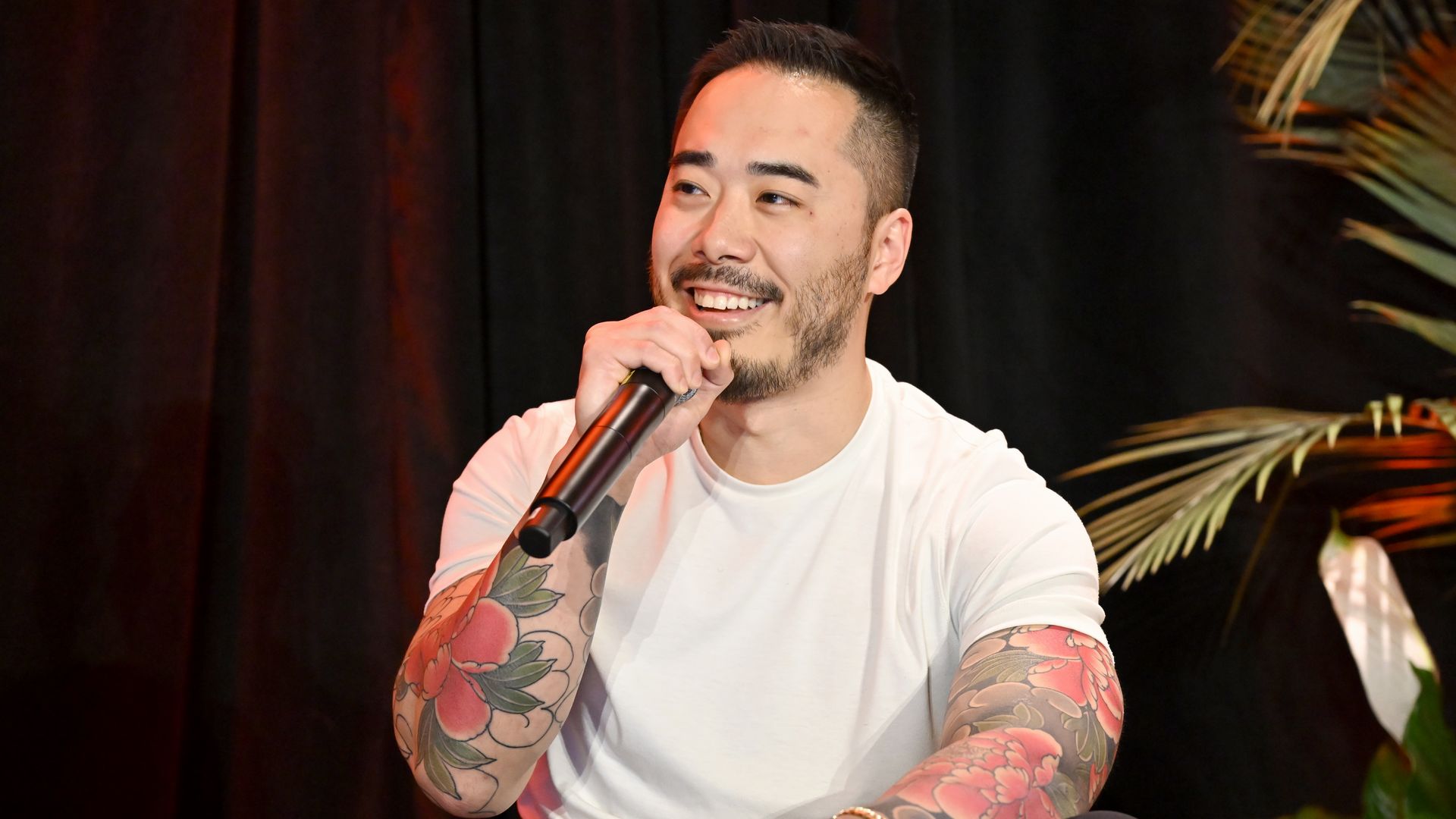 A man with a short beard and flower tattoos on his arms is holding a microphone and speaking in front of  curtain and plants. 