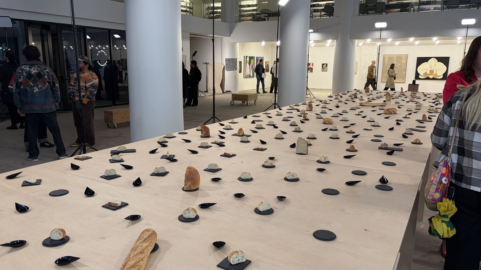 A modern gallery installation with a long white table covered in bread loaves and small black plates arranged in rows; visitors walk among white columns and framed artworks.
