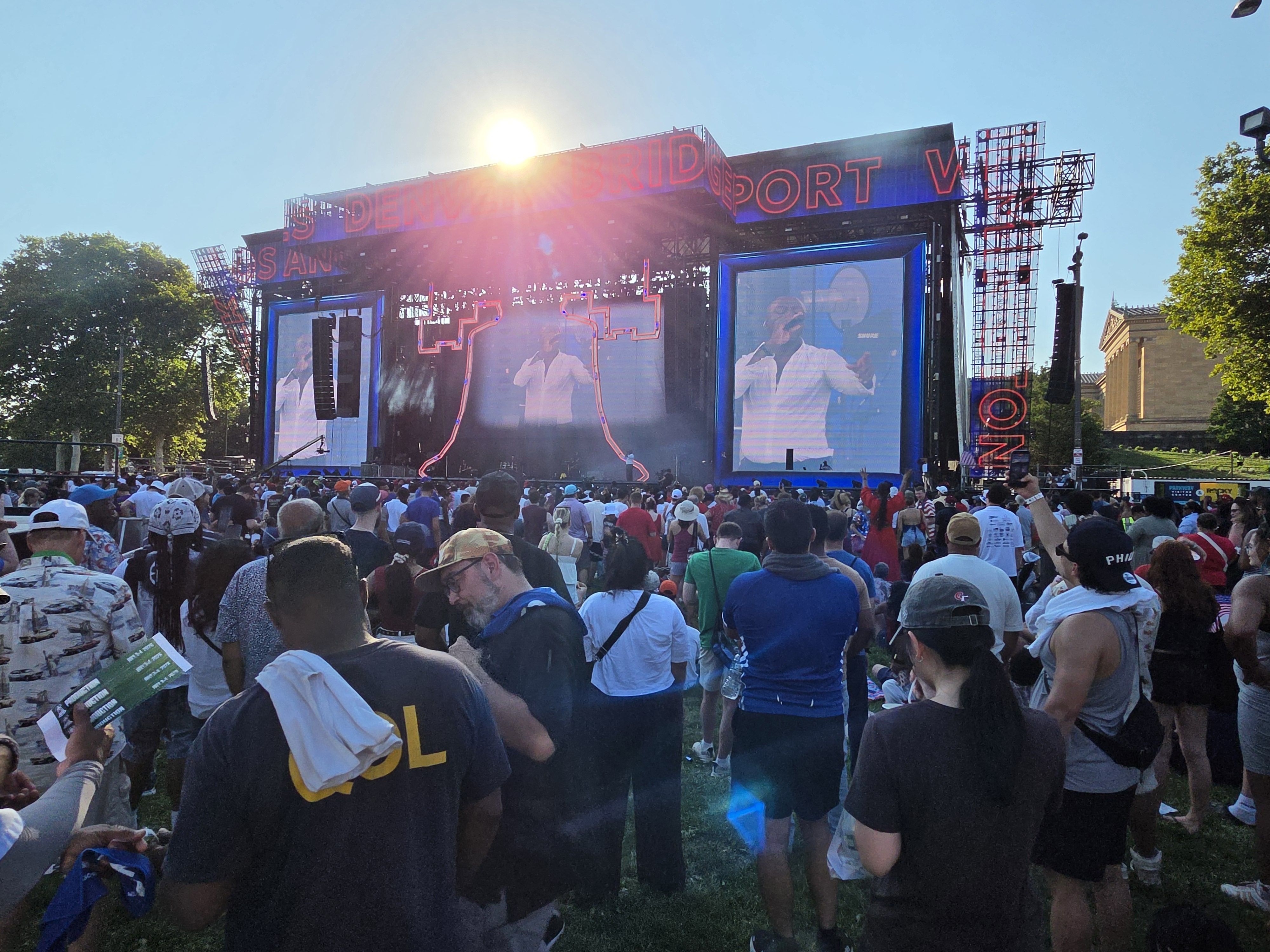 Outdoor music festival stage with bright neon signage and two large video screens showing performers; a dense crowd below, sun glare over the top, blue sky and trees on the sides.