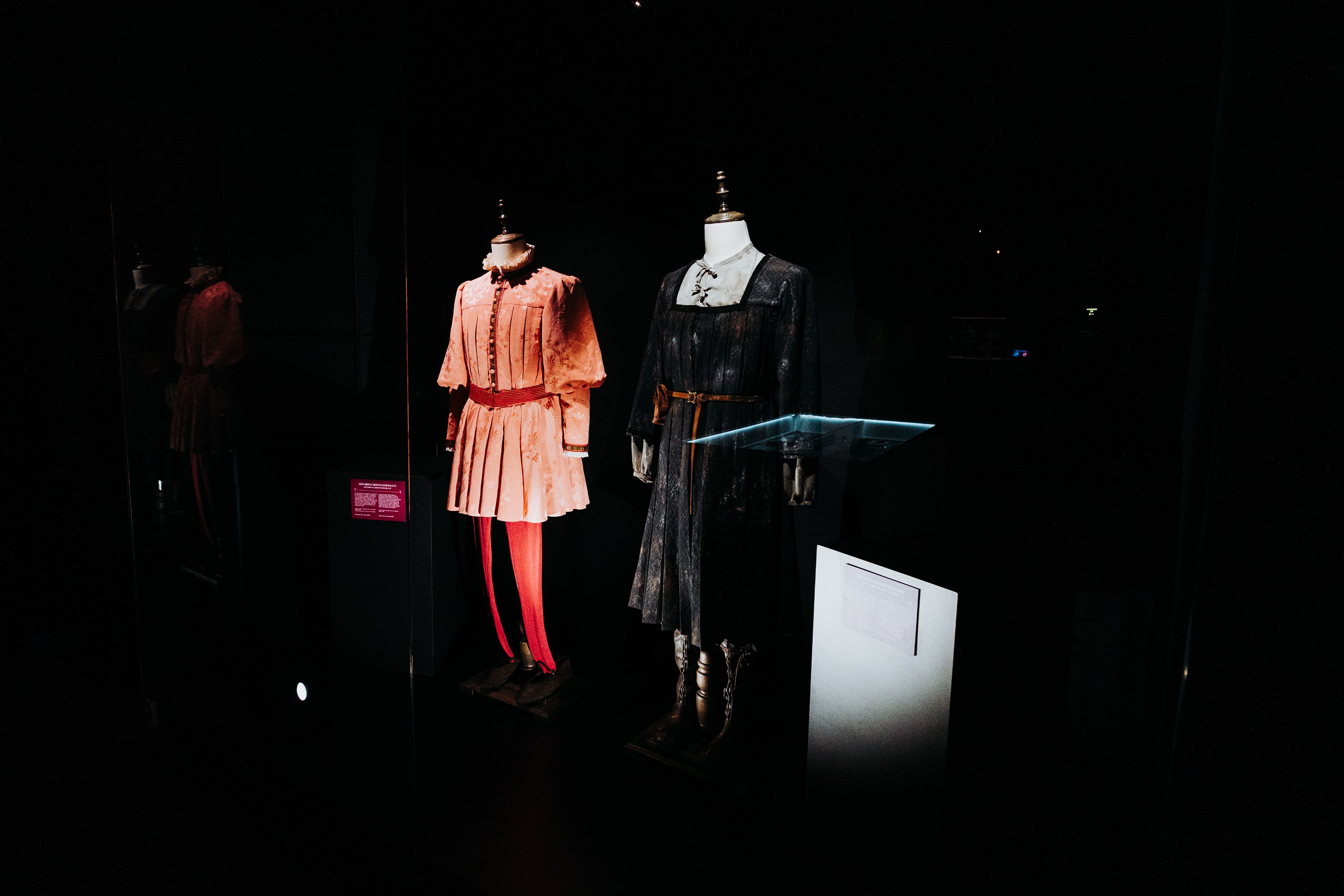 Dim museum display with two vintage dresses on mannequins: a pink pleated dress with a red belt, and a dark square-neck dress with a light gray bodice; glass cases nearby.