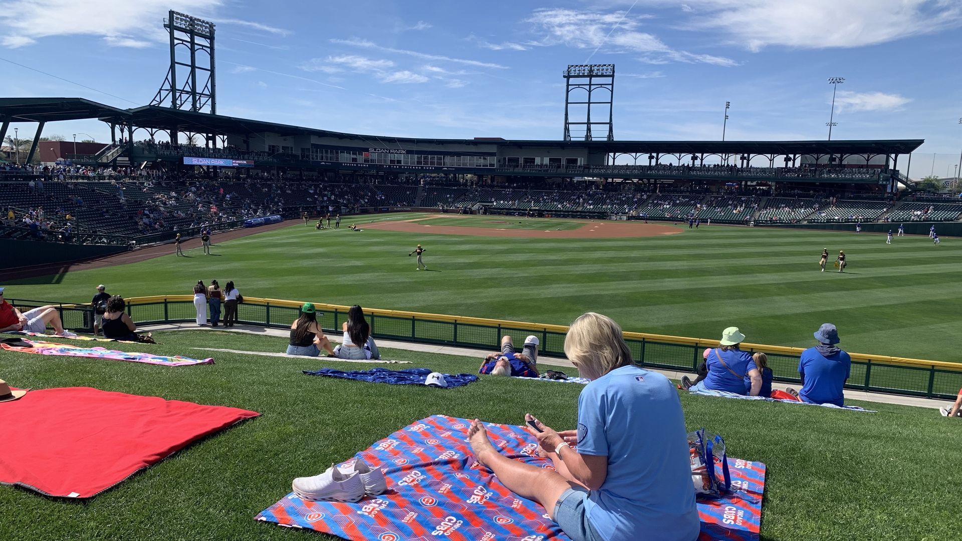 People sitting on blankets and grass hill watching a baseball game at Sloan Park under blue skies, some wearing Chicago Cubs colors and gear.