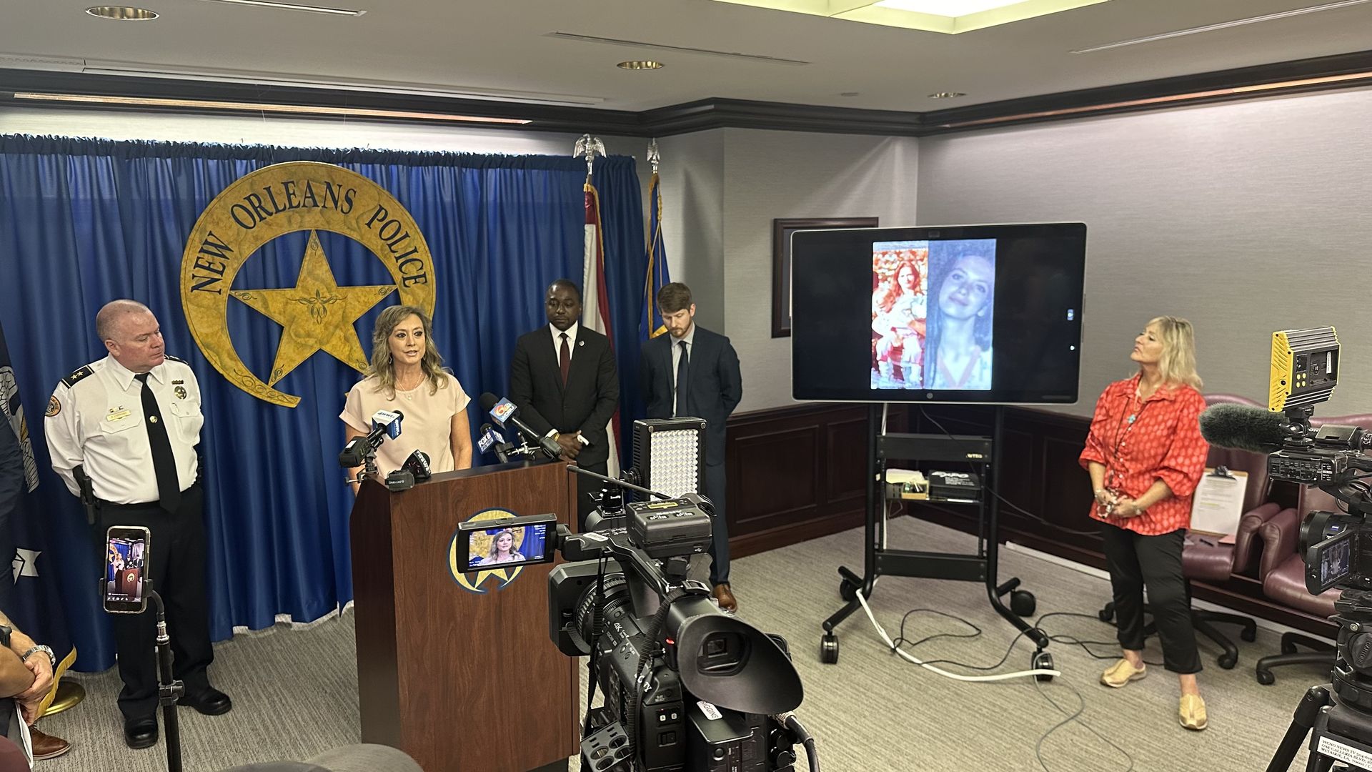 A woman stands at a podium as media cameras film her. The NOPD logo hangs on the wall behind her.