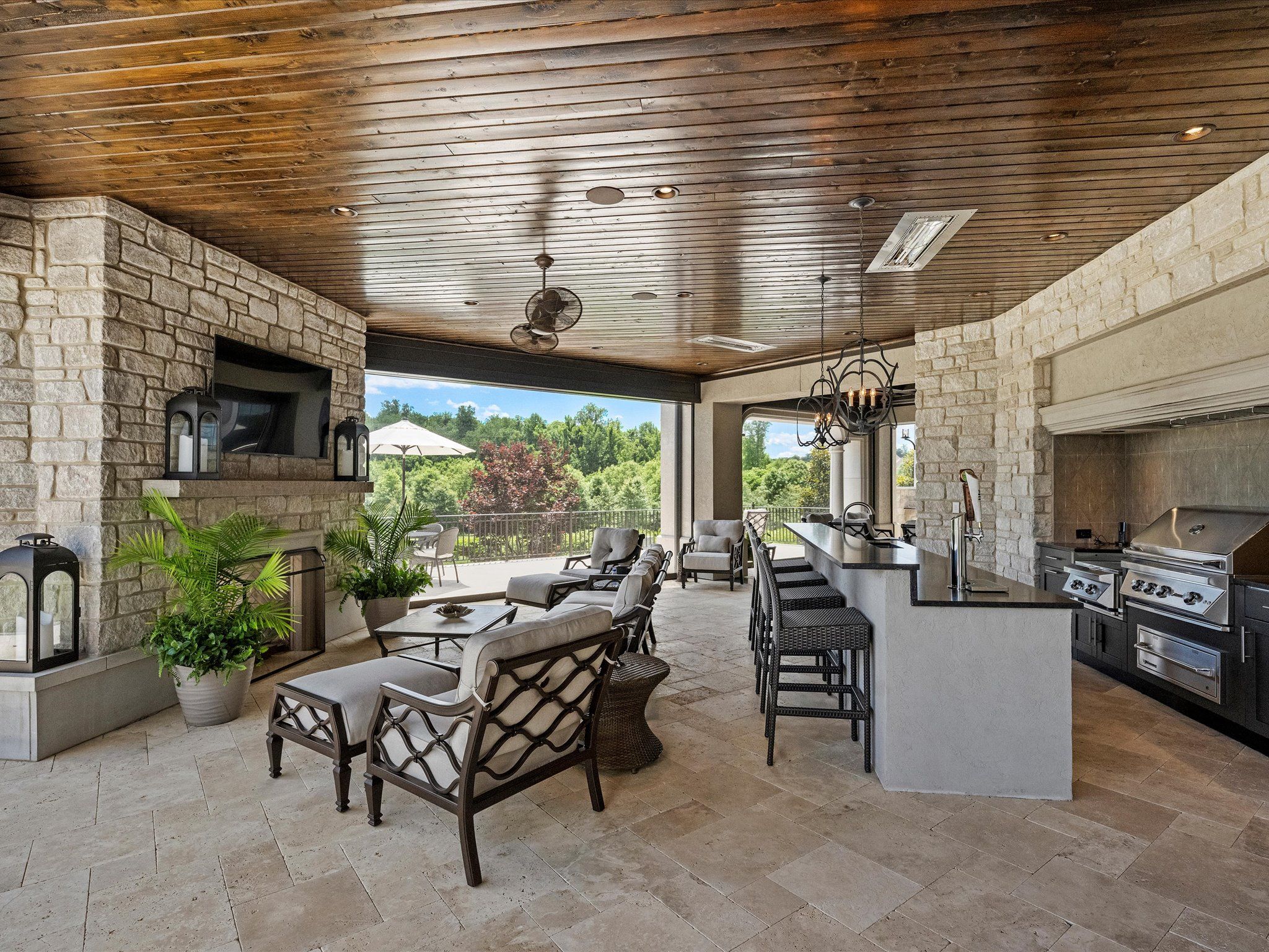 Spacious outdoor patio with stone walls, wooden ceiling, cushioned seating, a bar counter with stools, built-in grill, fireplace, and view of green trees and blue sky outside.
