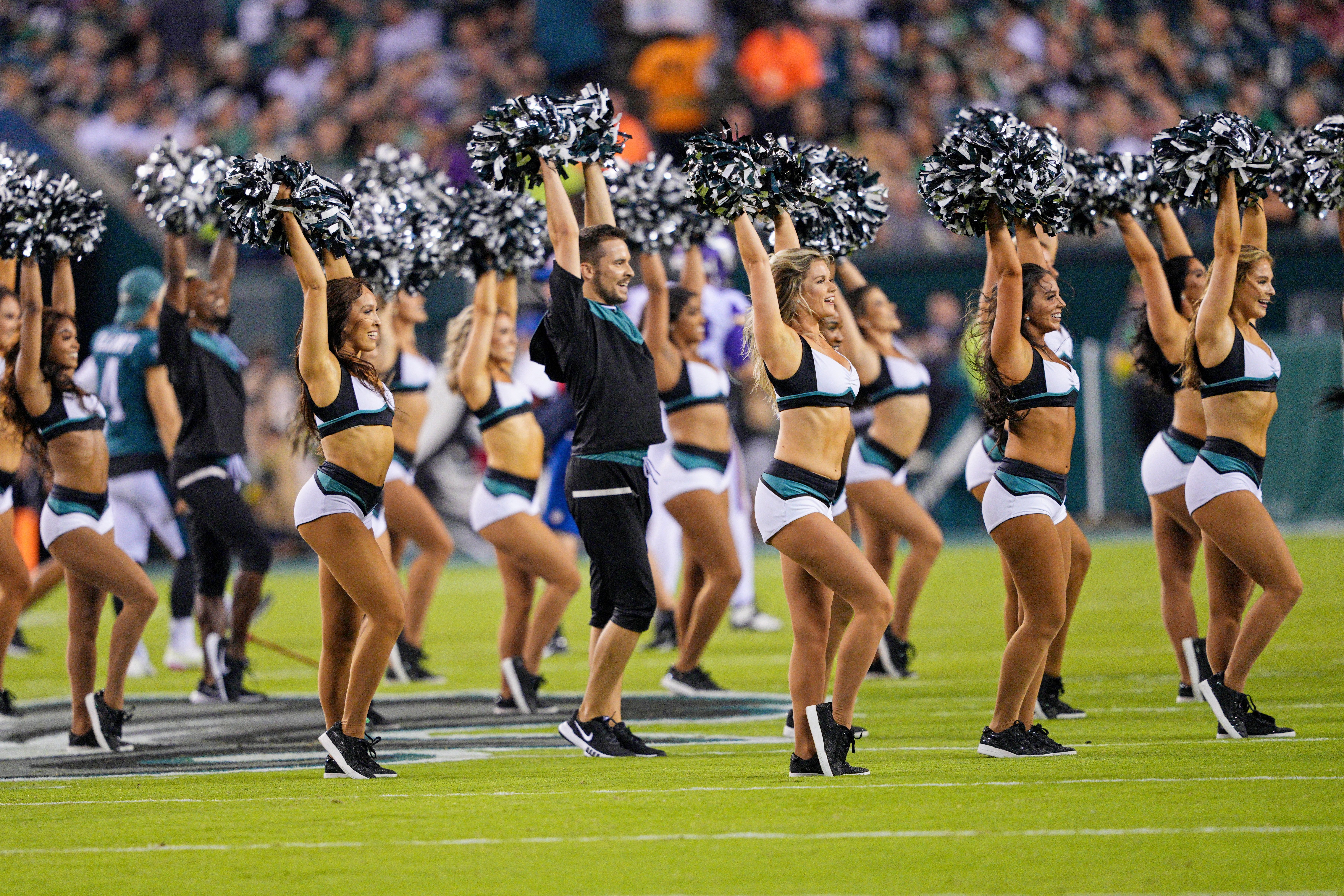 Cheerleaders and male dancers in black, white, and teal outfits performing on a football field, holding black and silver pom-poms, with a blurred crowd in the background.