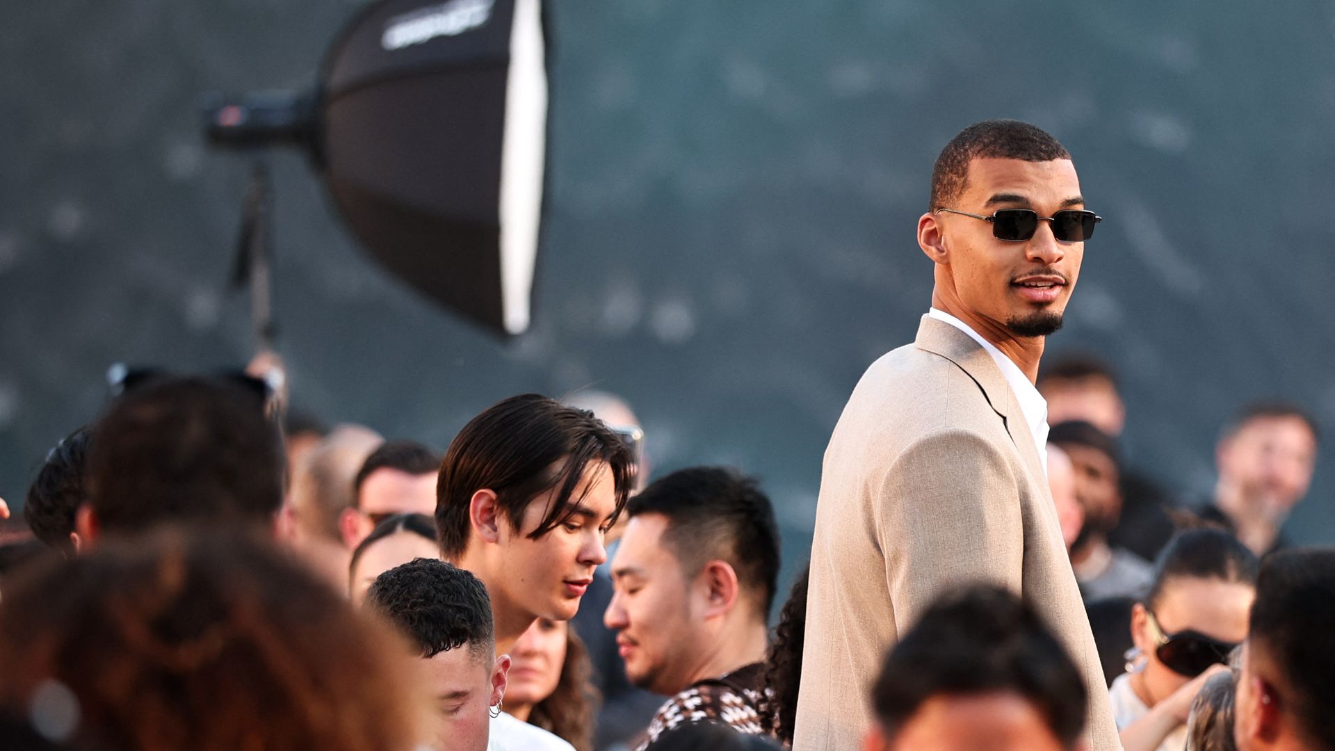 A man in a light beige suit and dark sunglasses stands among a crowd at an outdoor event, with a blurred photography softbox visible in the background.