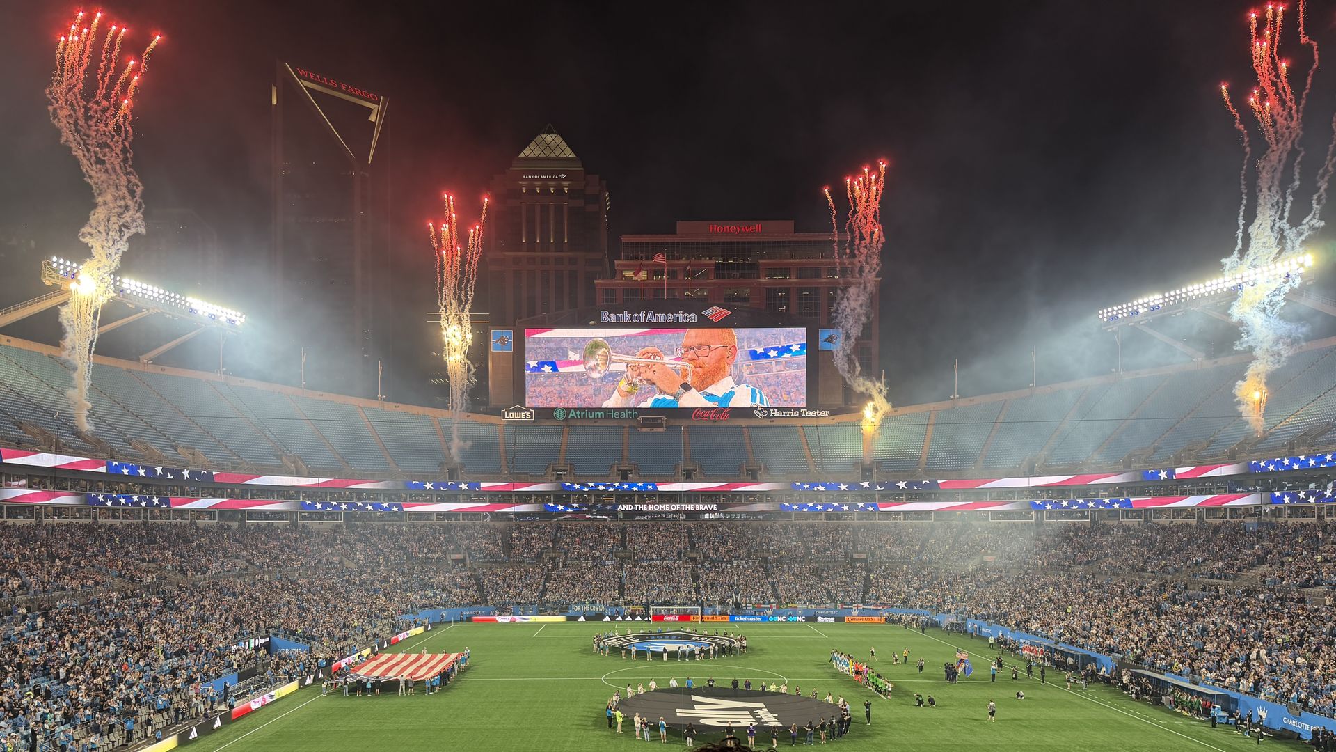 Nighttime at a packed football stadium as fireworks shoot above blue-seated stands. A large central screen glows, while fans fill the arena; a circular formation sits on the green field under red, white, and blue bunting.