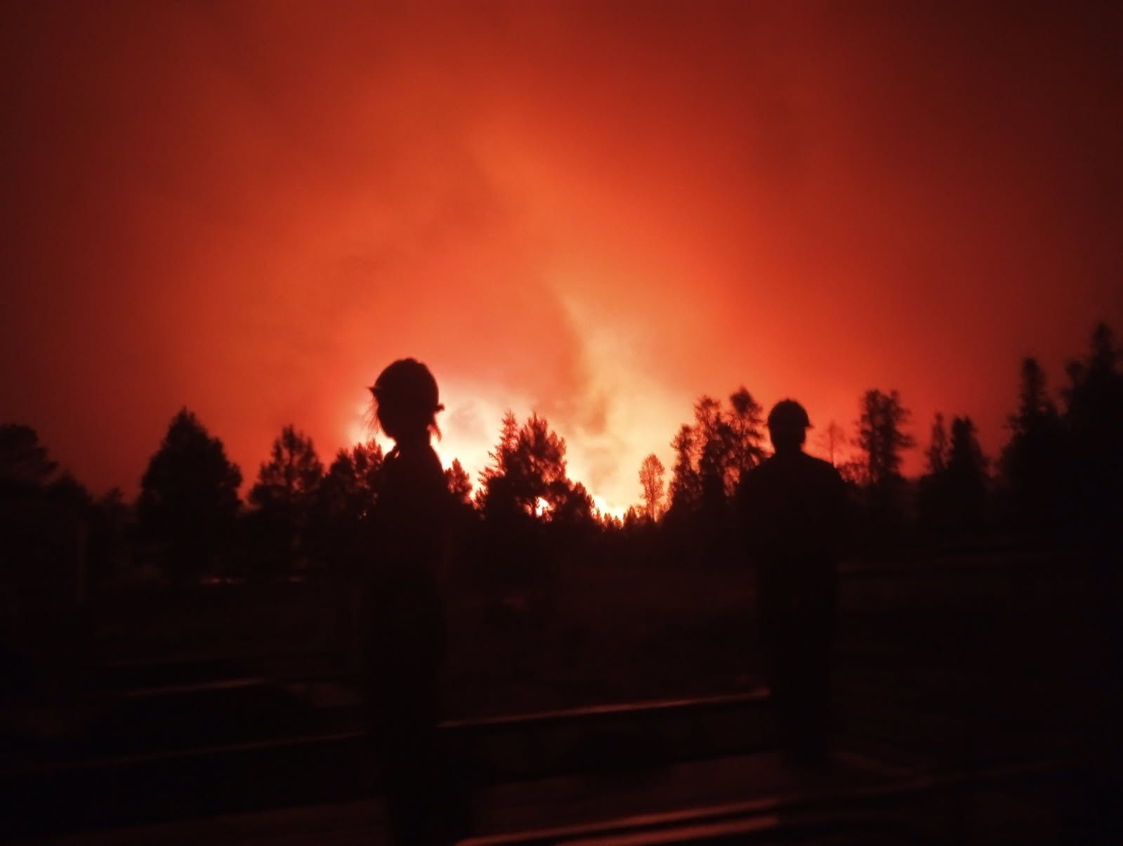 Two firefighters stand silhouetted against an intense orange-red sky as the Dragon Bravo Fire burns through a forested area at night, with flames and smoke rising in the background at Grand Canyon National Park on July 12, 2025.