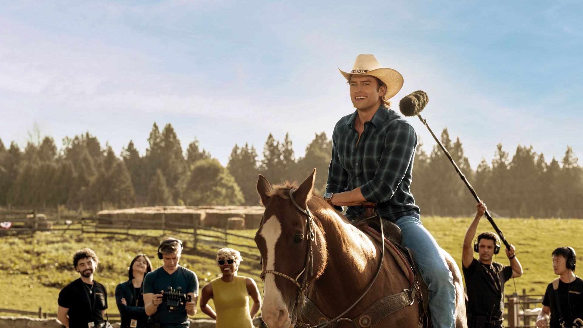An actor in a cowboy hat and plaid shirt rides a brown horse in a sunny pasture while a film crew with cameras and boom mic records him, with trees and fencing in the background.
