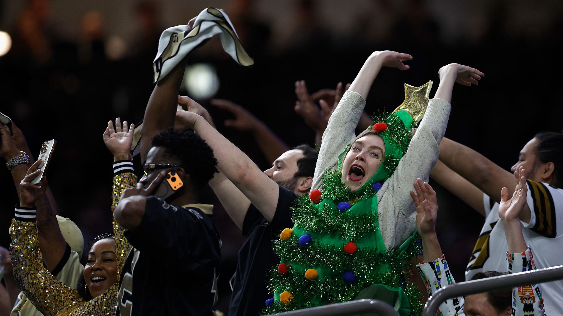 A person dressed as a Christmas tree celebrates in the stands at a stadium with people dressed in black and gold Saints jerseys and outfits.