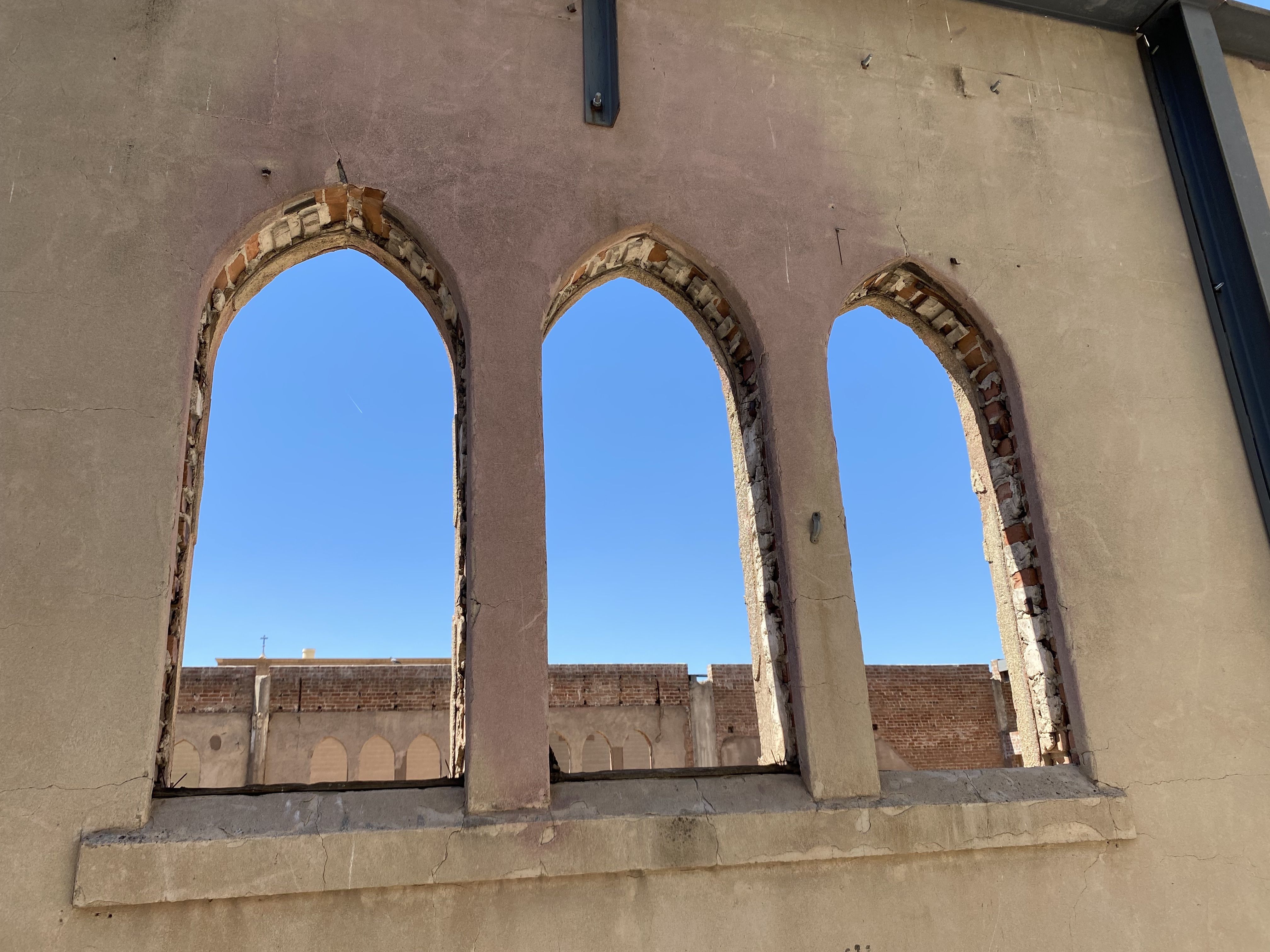 Three tall windows with no glass with rose colored tinting on the beige walls. 