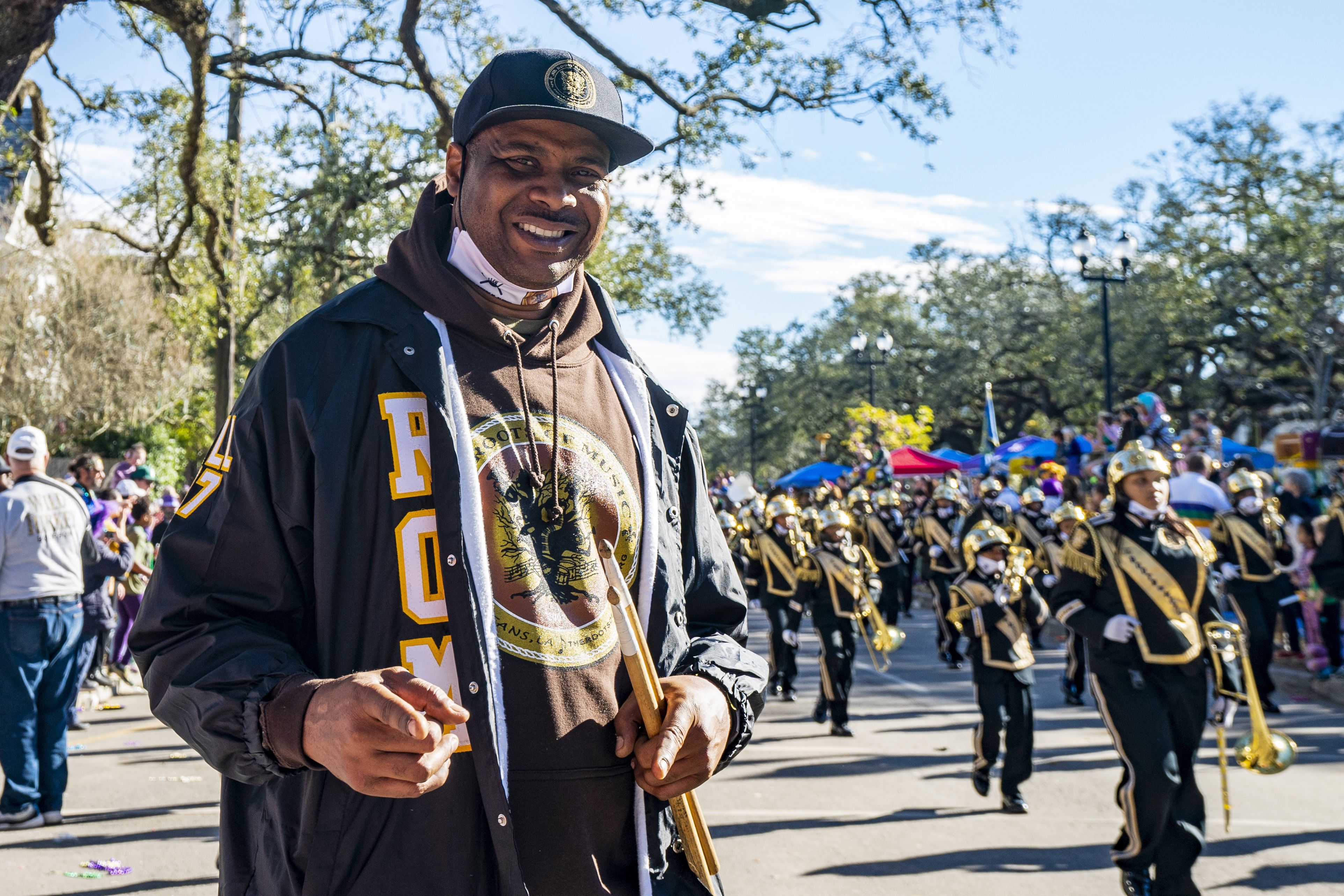A man smiles toward the camera as a marching band processes during a Mardi Gras parade behind him.