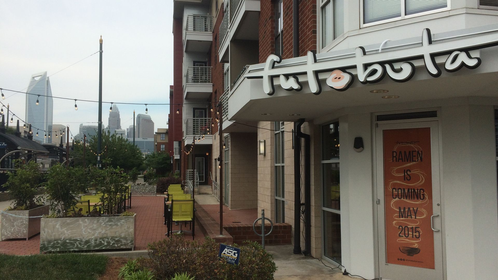Street view of futobuta ramen restaurant with orange sign on door saying 'RAMEN IS COMING MAY 2015', outdoor seating, string lights, and city skyline in background.