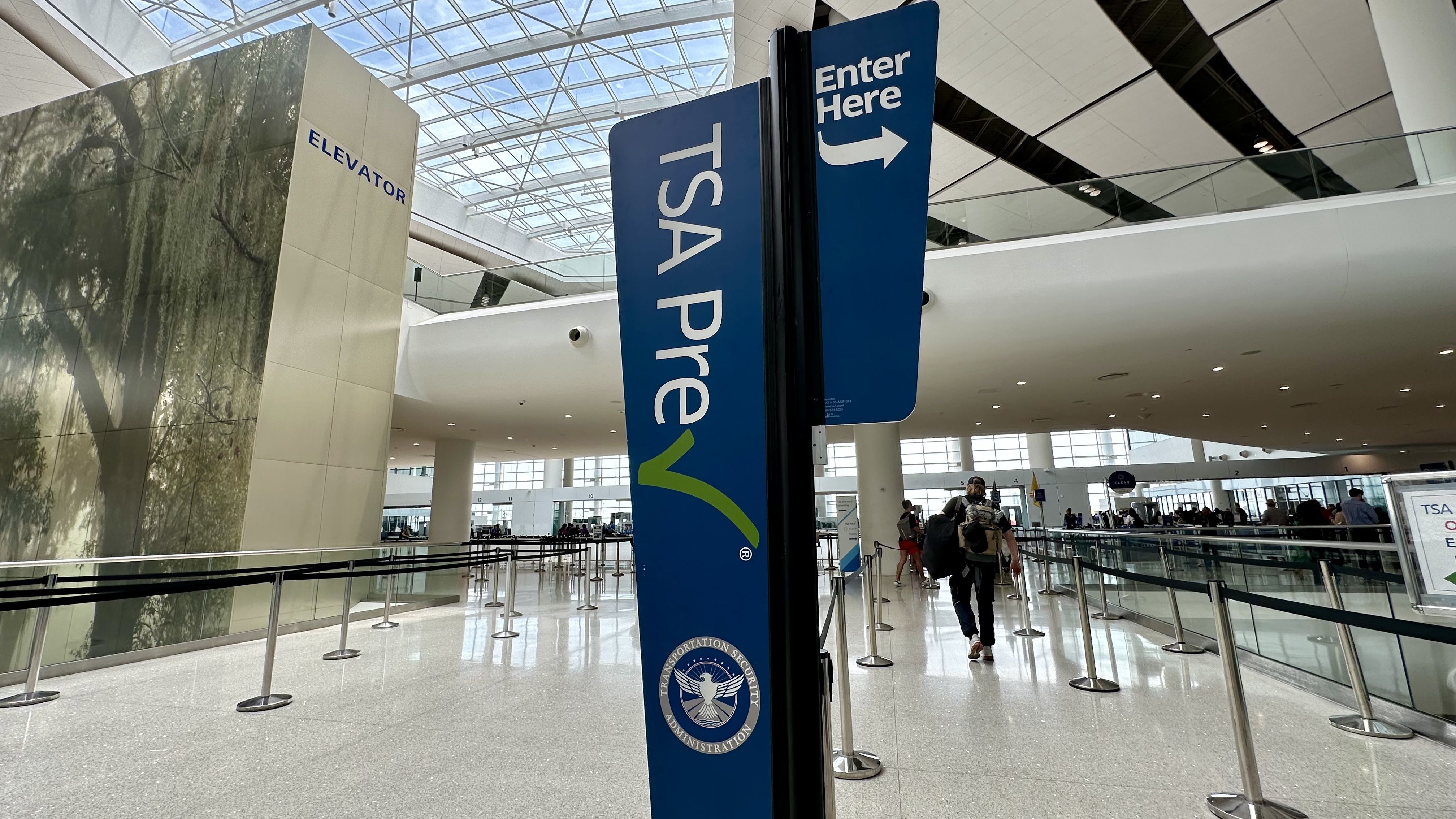 Bright airport terminal with a tall blue TSA Pre sign showing "TSA PRE" and an "Enter Here" arrow. A wall to the left reads "ELEVATOR"; security queue barriers and travelers beneath a glass skylight atrium.