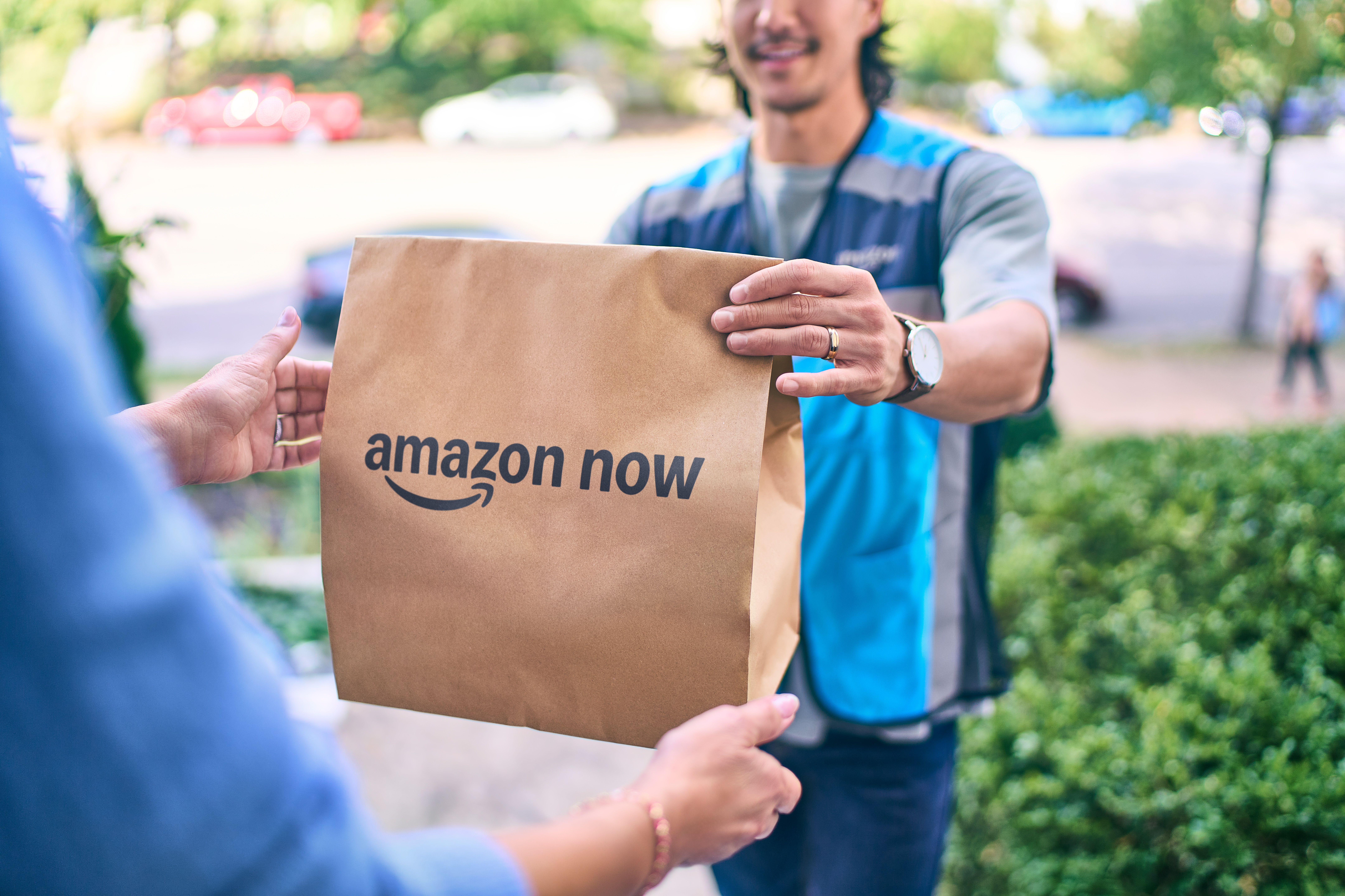 Delivery worker in a blue vest hands a brown paper bag labeled amazon now to a customer outside, with green trees and a street blurred in the background.