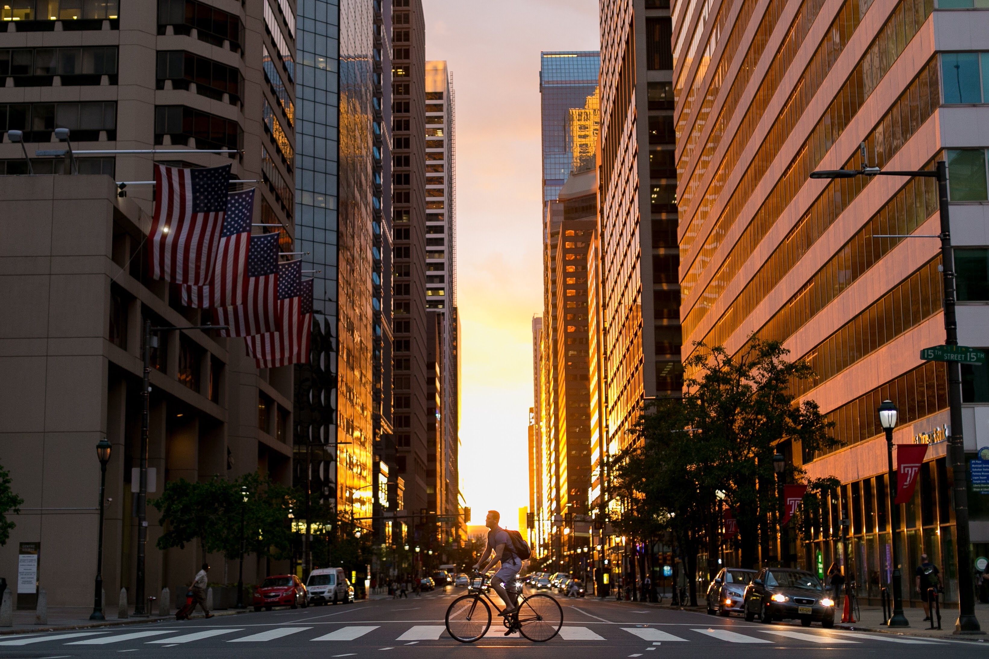 Sunset city street framed by tall buildings; a cyclist rides across a crosswalk as warm orange light washes the facades; American flags hang from a left building, with cars and pedestrians below.