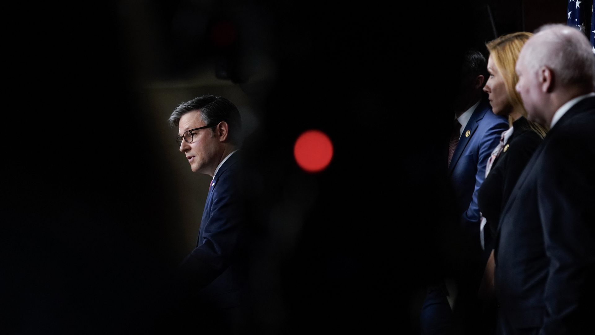 House Speaker Mike Johnson, seen through a silhouette, wearing a blue suit jacket and glasses, speaks to reporters in the Capitol's studio A flanked by fellow House Republicans.