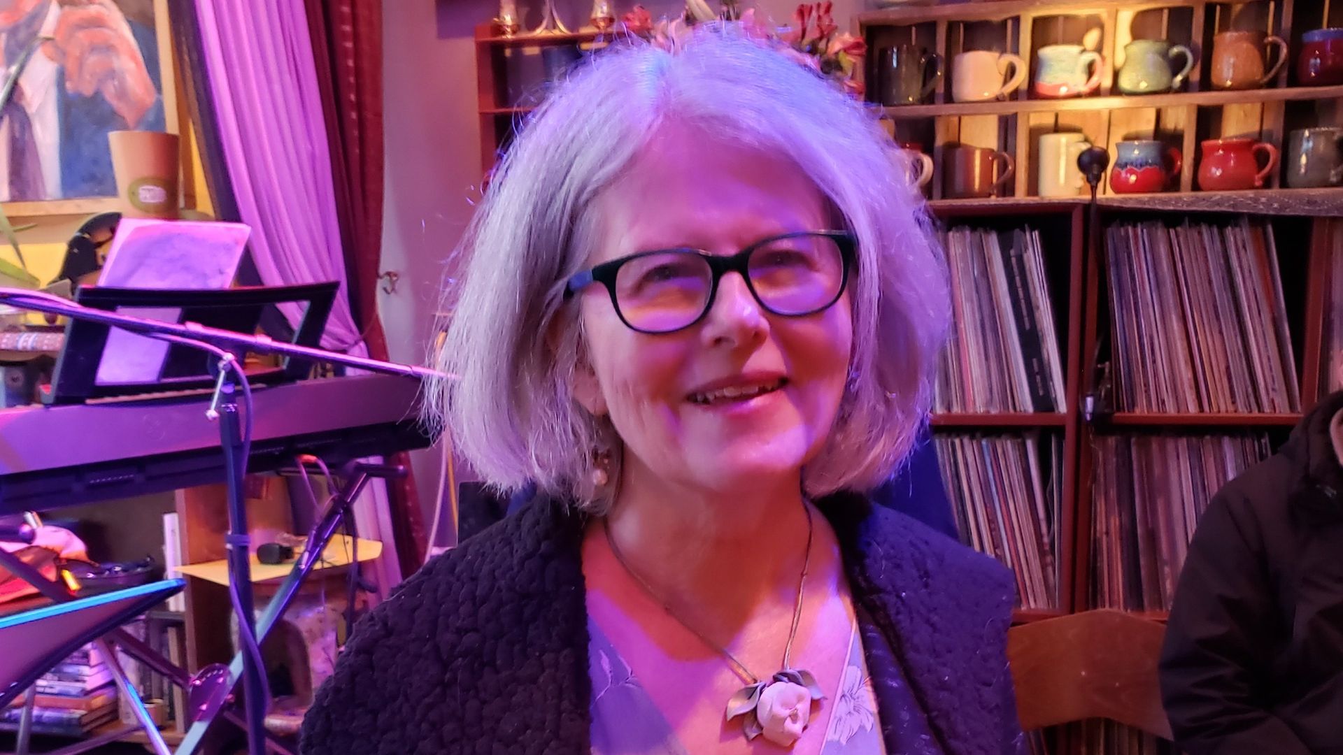 Woman with gray hair, glasses, smiling in front of shelves of records and a piano.