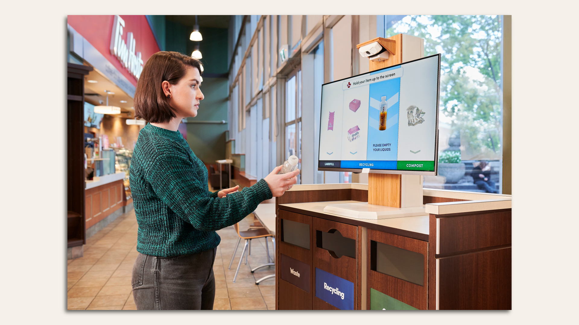 A woman faces a screen and a trash receptacle and views the screen to see which bin she should use for the item she's holding.