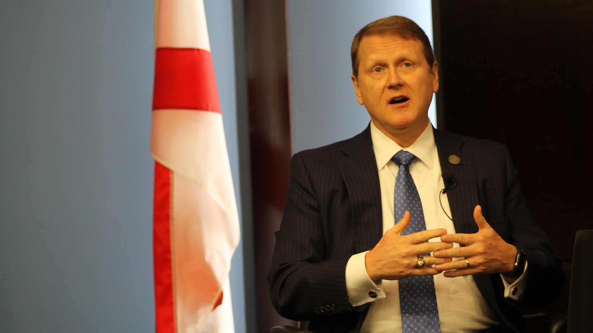 Man in a dark pinstripe suit and blue polka dot tie speaking with hands gesturing. A white flag with a red cross is partially visible to his left.