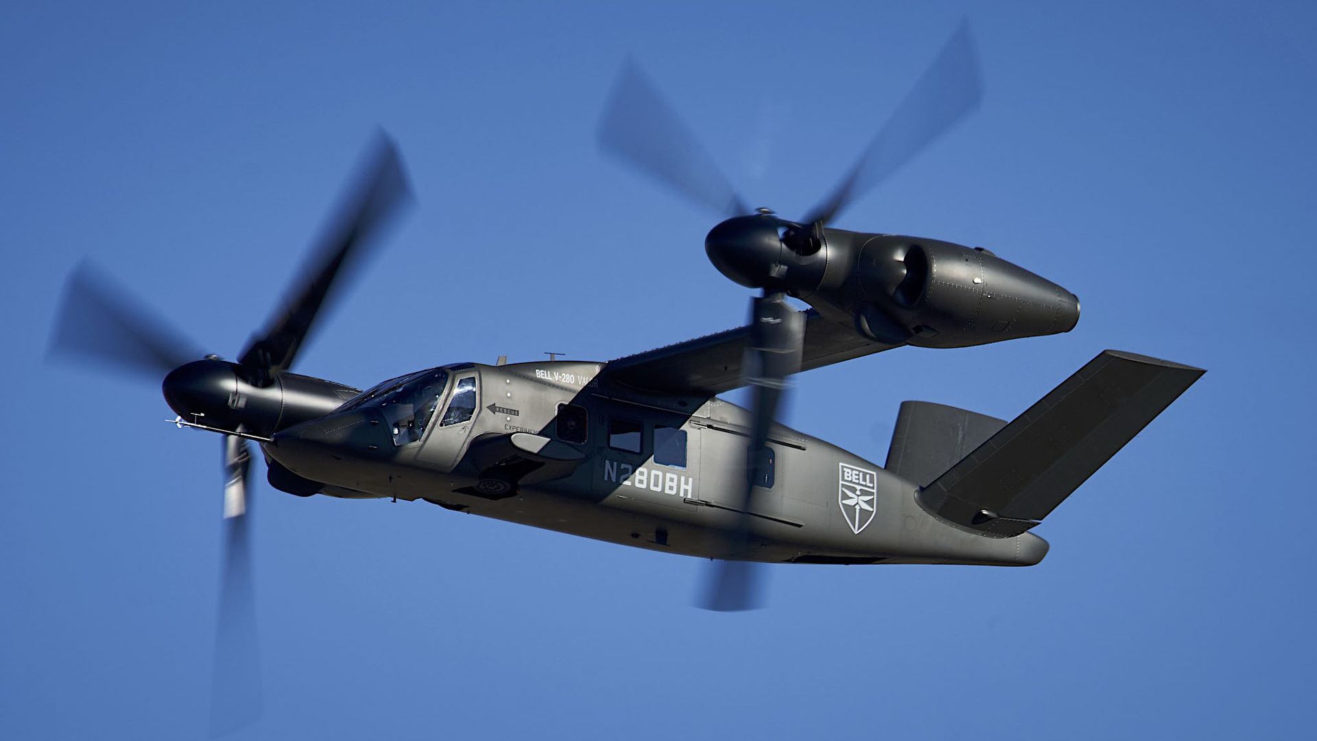 Grey Bell V-280 Valor tiltrotor aircraft in flight with spinning propellers against a clear blue sky.