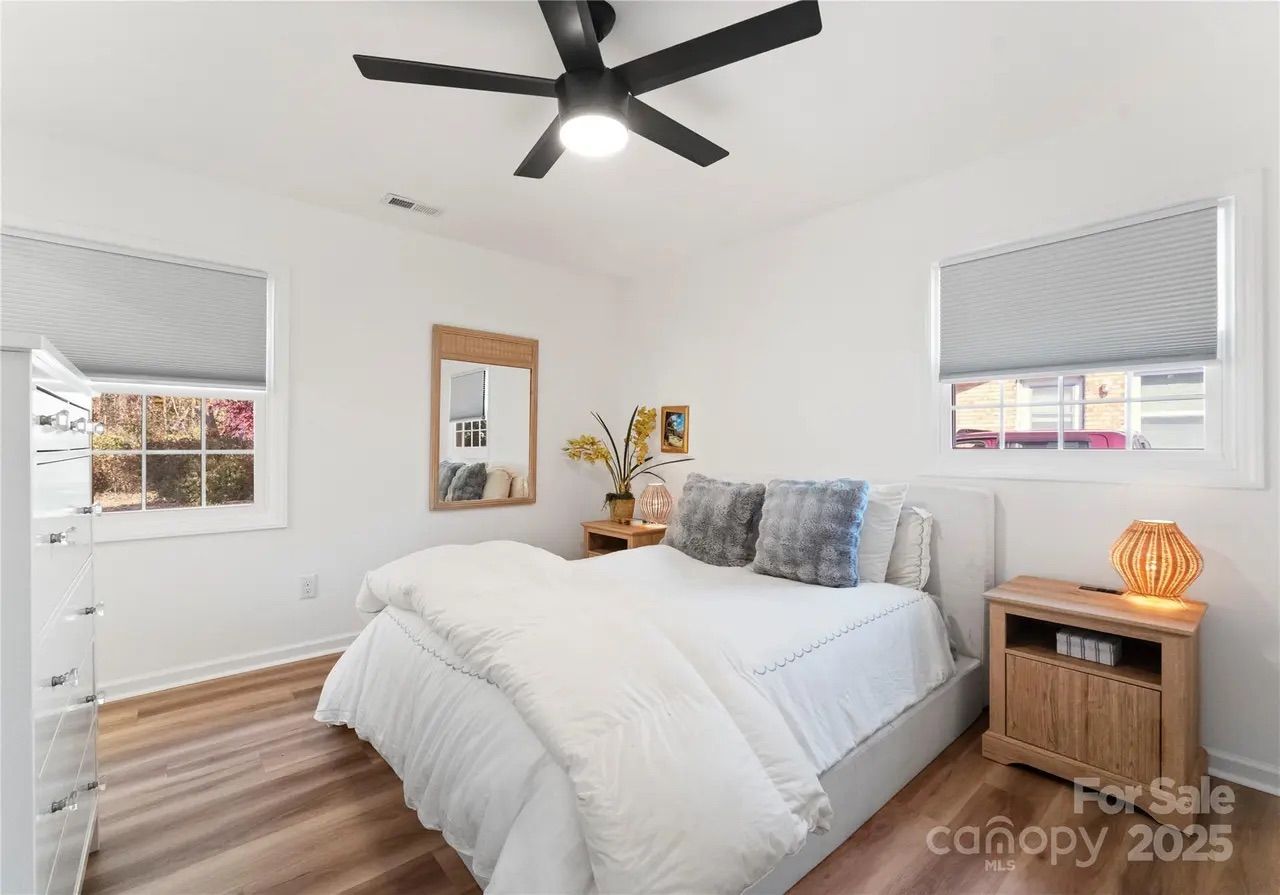 Bright minimalist bedroom with white walls, wood floor, white bed with gray pillows, two wooden nightstands with lamps, a wall mirror, two windows with gray blinds, and a black ceiling fan.