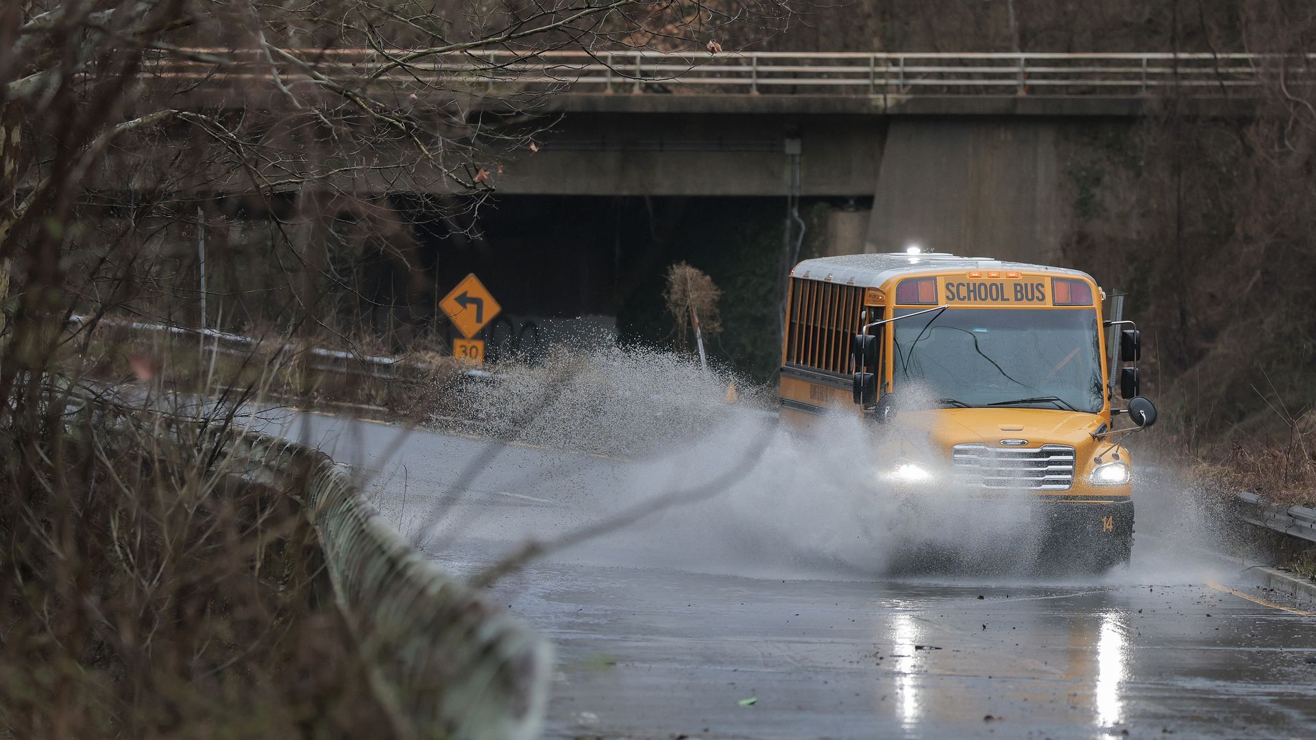 Yellow school bus splashes through a flooded wet road under a bridge; headlights on, an orange SCHOOL BUS sign visible, with a guardrail and a left-turn warning sign in the background.