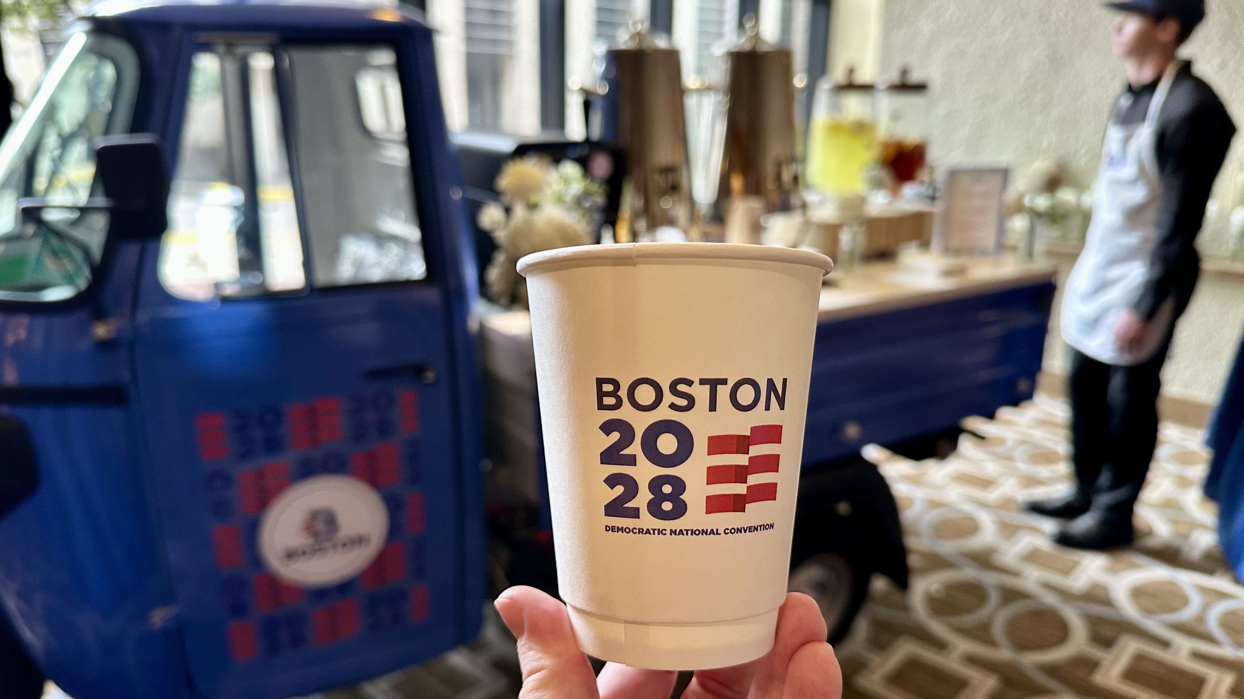 Paper cup in foreground branded with "BOSTON 2028" and "DEMOCRATIC NATIONAL CONVENTION", held by a hand. Blurred background shows a blue vehicle/food cart and a vendor in an apron at a beverage station.
