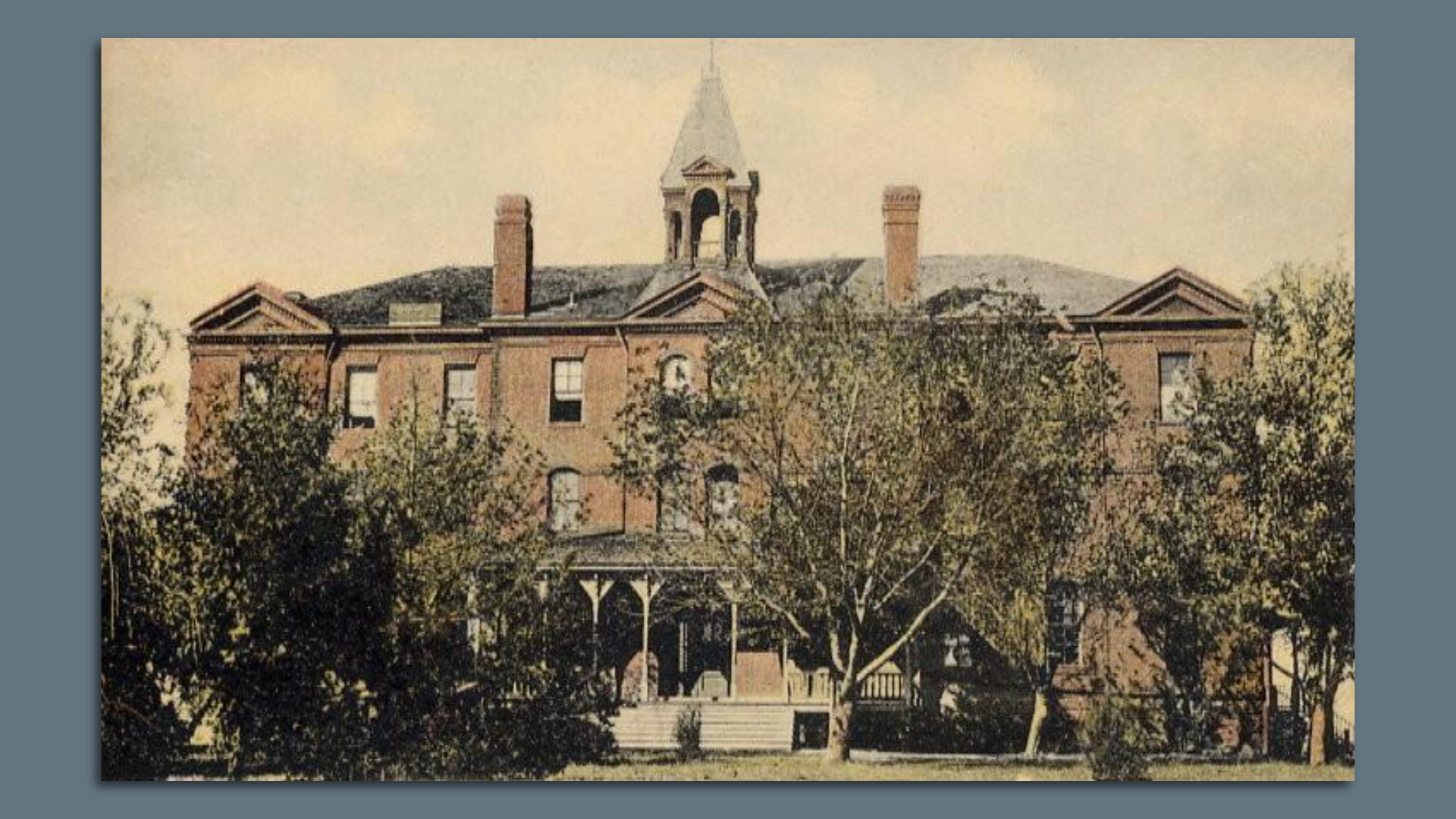 A large 19th century brick building with a bell tower and trees in front.