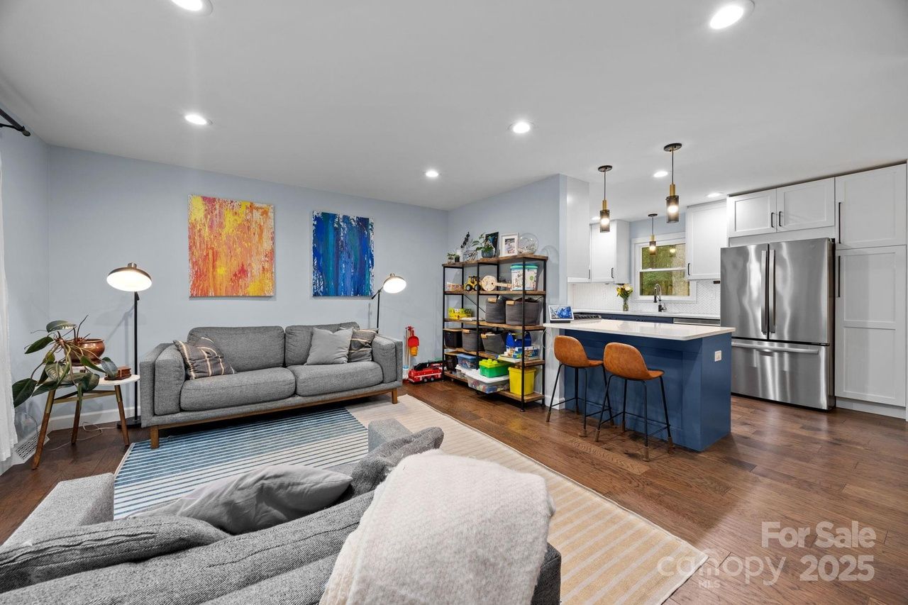 Open living room and kitchen with gray sofas, colorful abstract paintings, striped rug, blue kitchen island with brown bar stools, white cabinets, and stainless steel fridge.