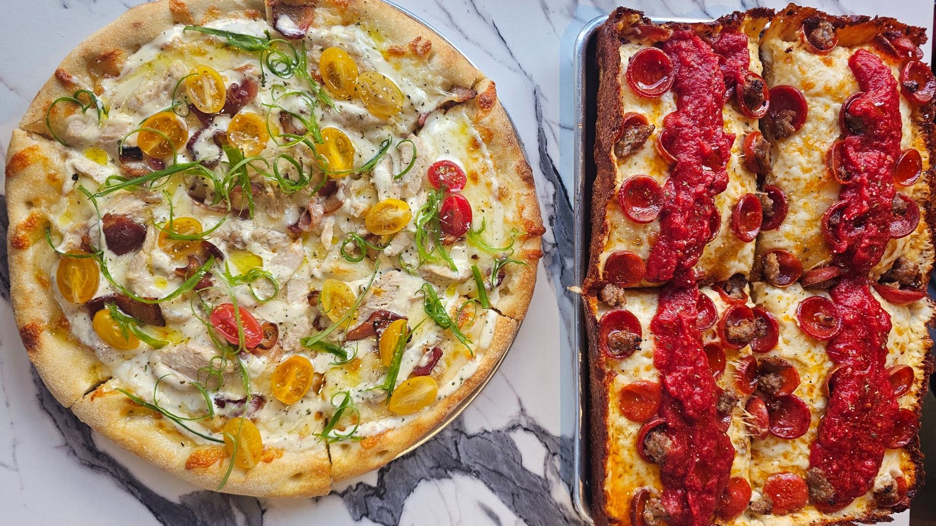 Two pizzas on marble surface: a round white sauce pizza with yellow and red cherry tomatoes, green onions, chicken, and bacon on the left; a rectangular deep-dish pepperoni pizza with sausage and tomato sauce on the right.