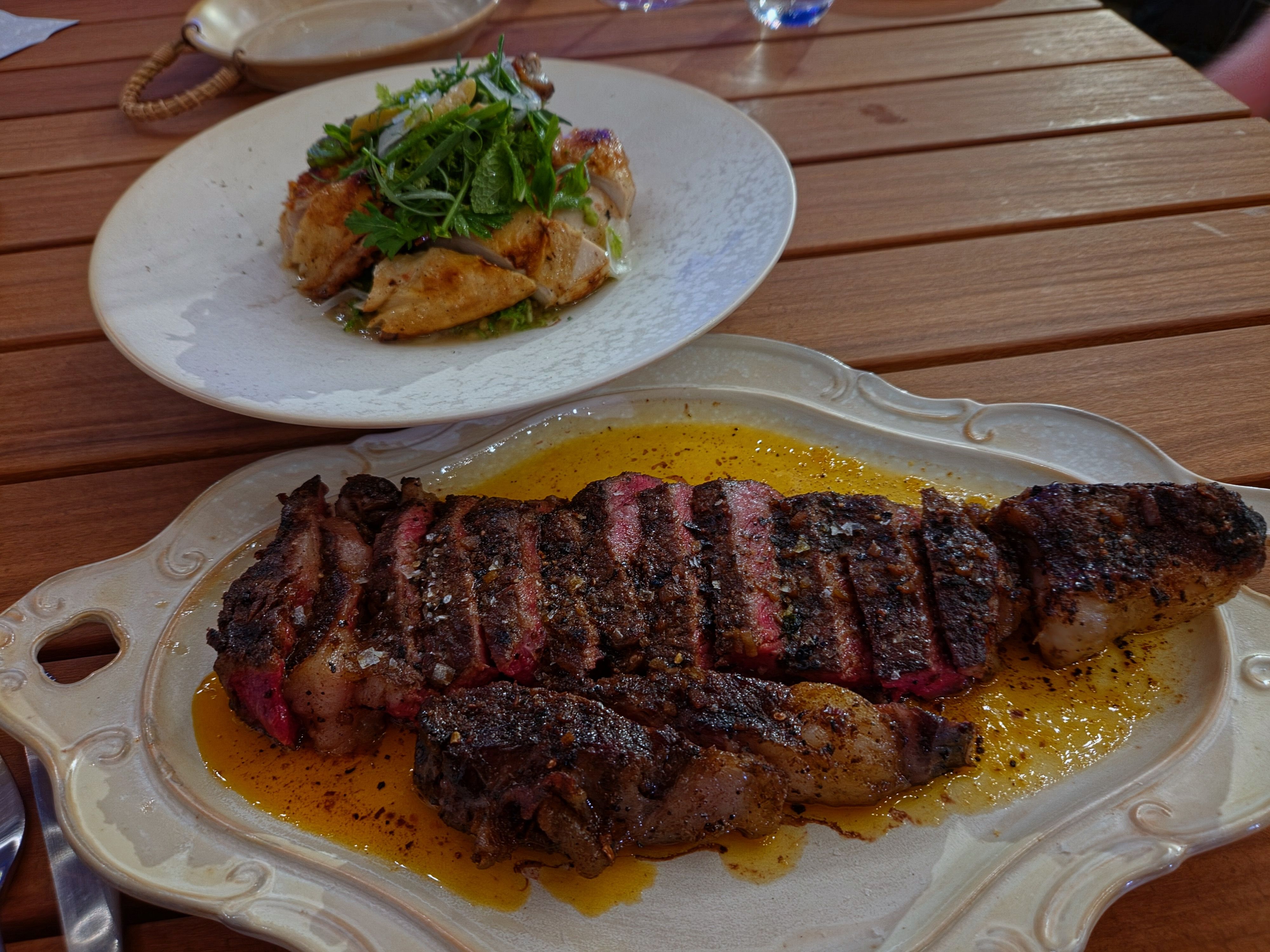 Two plates on a wooden table: a long platter of pepper-crusted beef ribs in yellow sauce, and a white plate piled with fresh herbs and greens.
