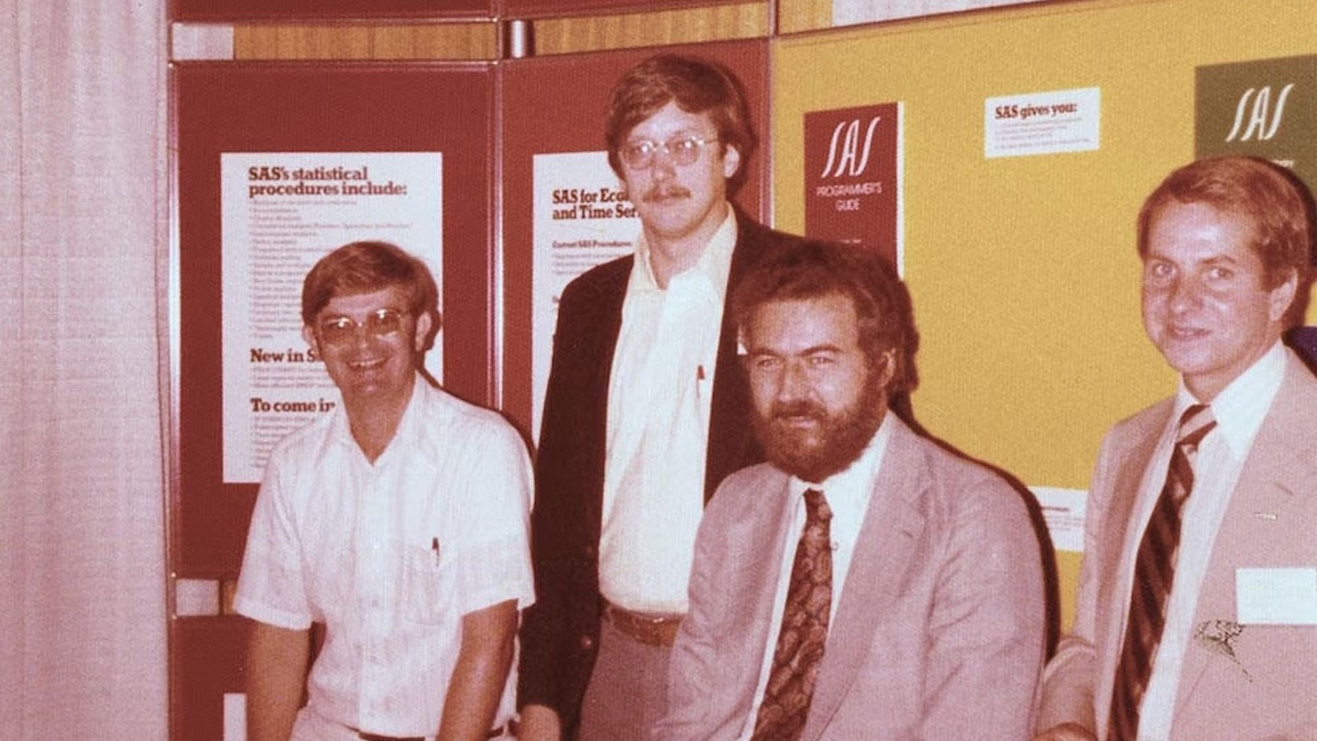 Four men in business attire pose at a SAS software booth with a yellow backdrop, posters about SAS procedures, and a banner reading "a new concept in software".