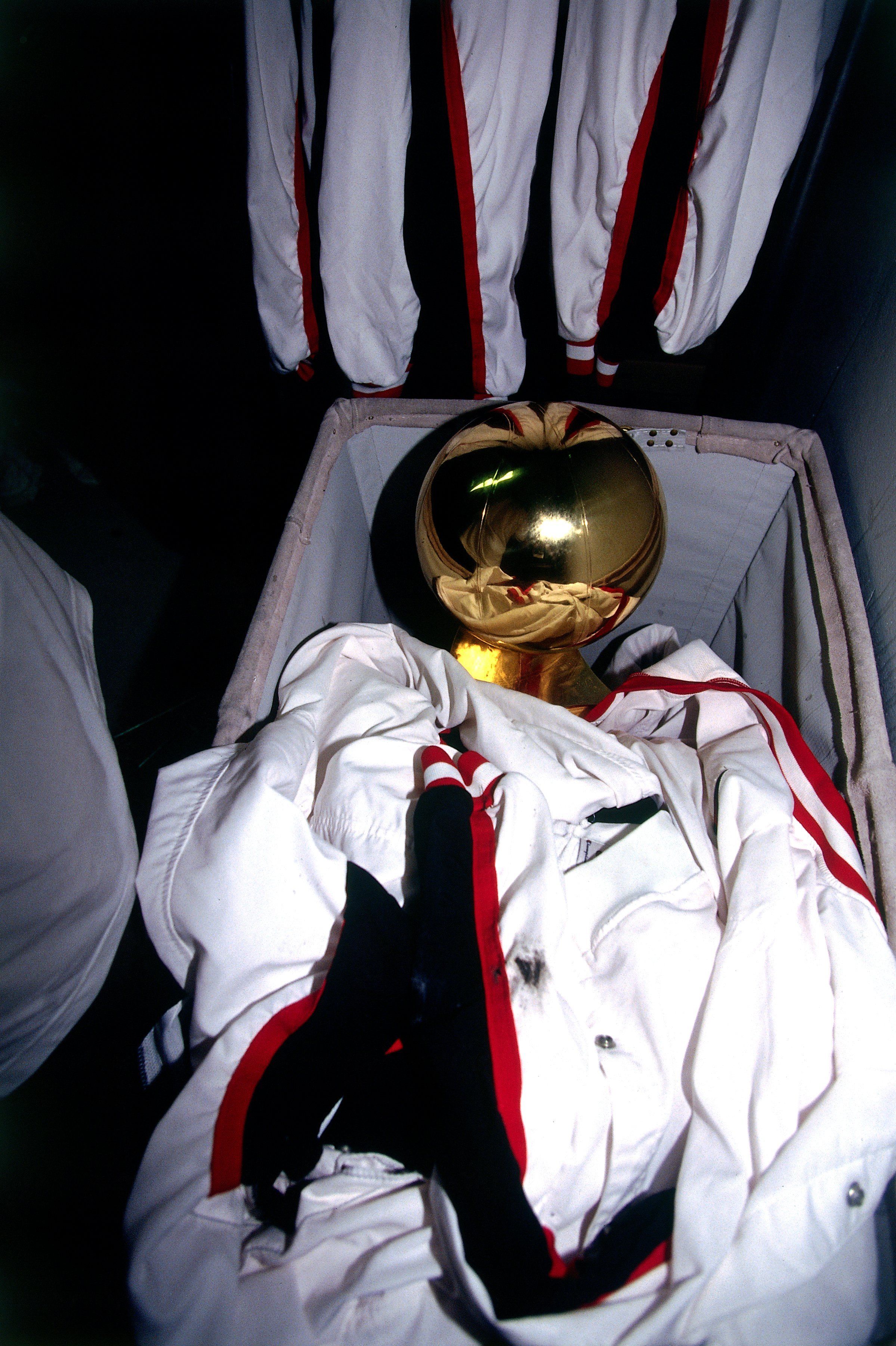 Shiny gold helmet rests inside a padded case with a white spacesuit and red trim, while hanging white suits with red edges loom in a dark closet-like space.
