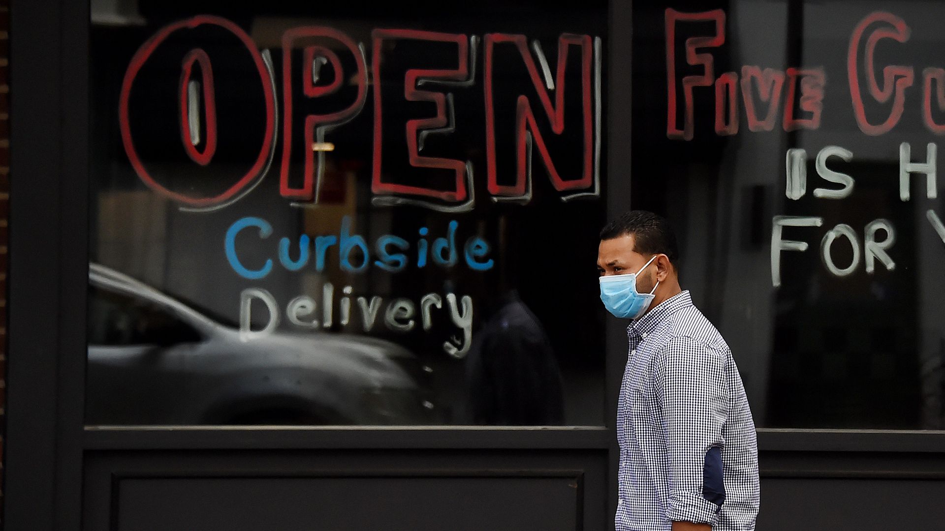 In this image, a man wears a mask and stands in front of a sign that reads "Open curbside delivery"