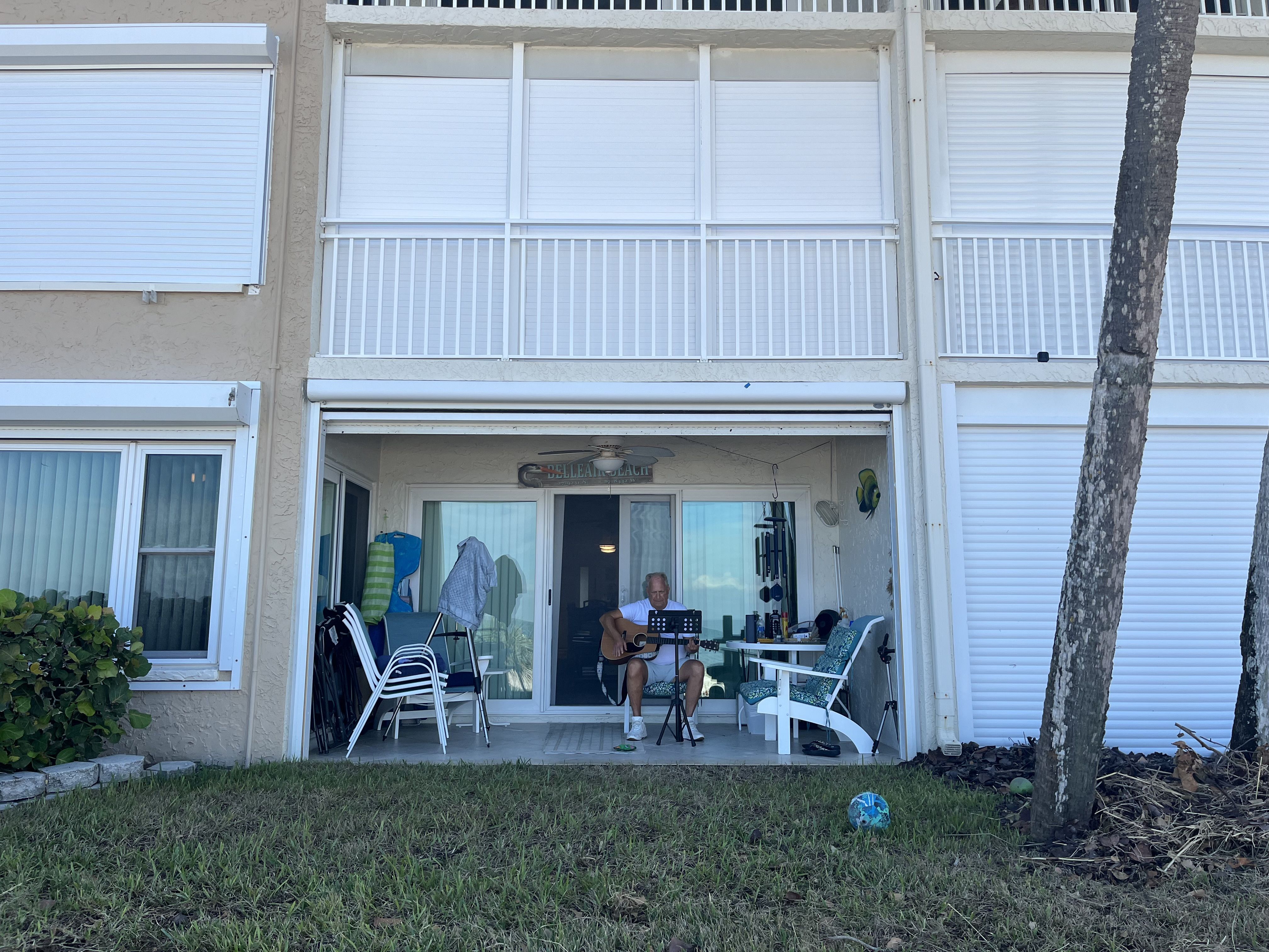 A man in a white T-shirt strums an acoustic guitar on a patio, surrounded by Adirondack chairs.