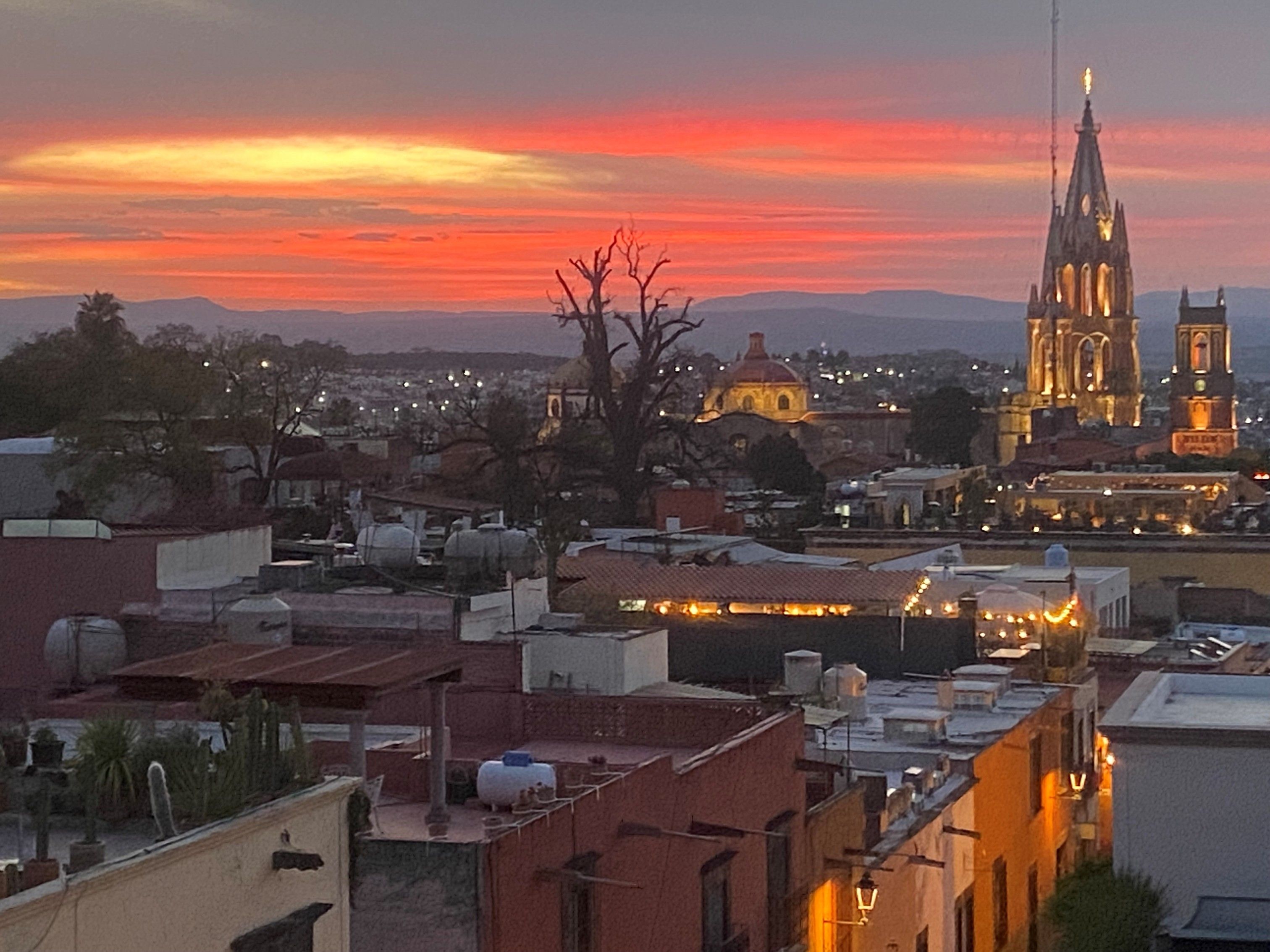 Sunset over a cityscape with orange and purple skies, illuminated historic buildings including a tall church tower, rooftop water tanks, and scattered trees.