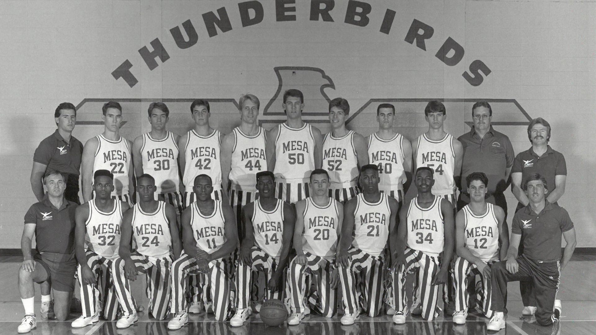 Black-and-white portrait of the Mesa Thunderbirds basketball team in uniforms with MESA on jerseys and striped pants, flanked by coaches under a large THUNDERBIRDS arch; basketball at their feet.