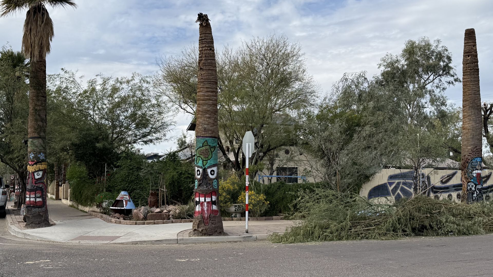 Three tall palm trees with colorful tribal face paintings on their trunks standing near a sidewalk and street corner, with surrounding greenery and a cloudy sky.