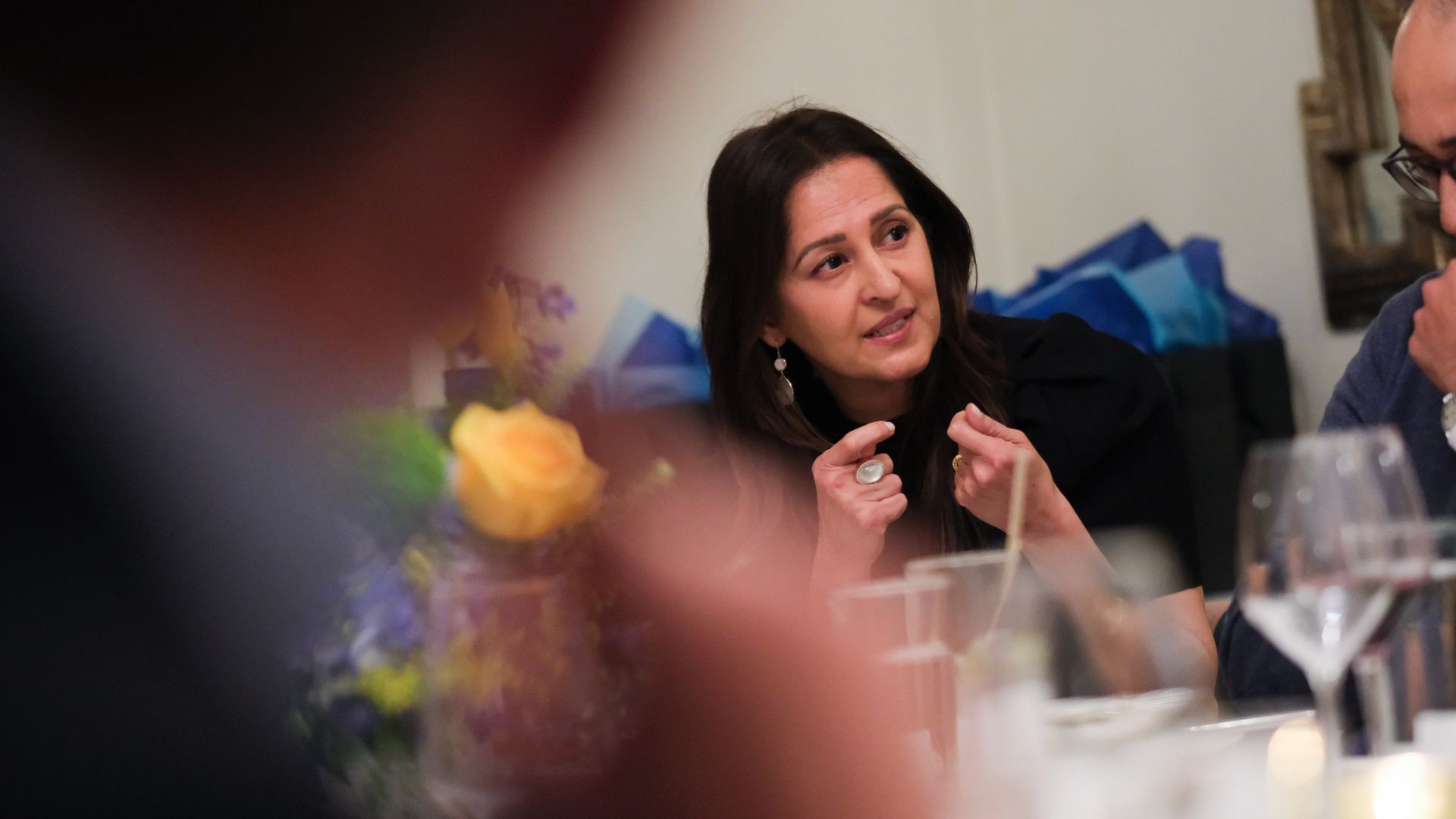Center: a woman with dark hair wearing pearl earrings speaks, hands clasped, at a dinner table. Blurred foreground, background shows blue cushions and a man wearing glasses nearby.