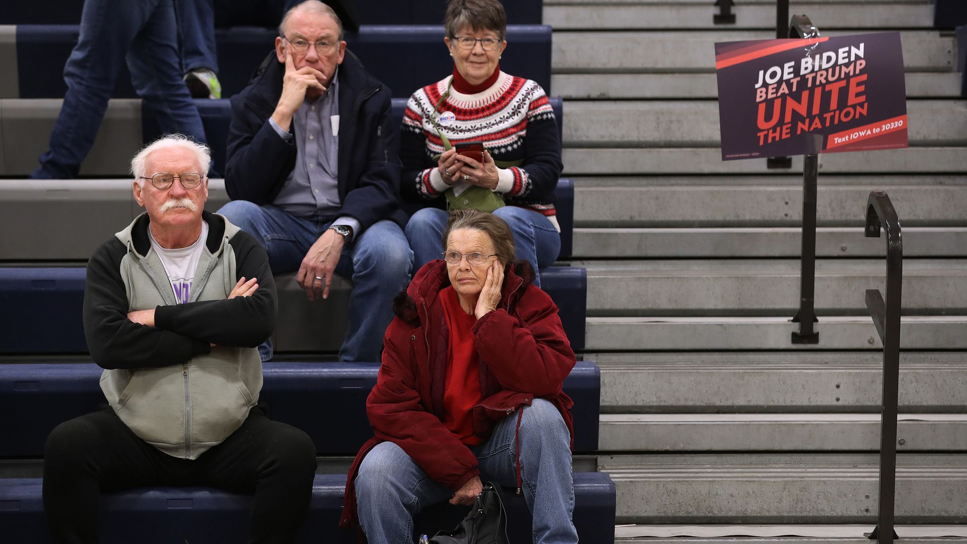 Photo of caucus participants looking glum sitting on bleachers next to a Biden sign