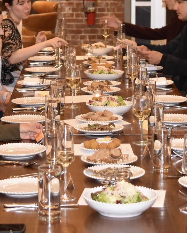 Long wooden table with diners on both sides, white bowls of salad and breaded appetizers, and tall wine glasses. A brick wall and fire extinguisher are visible in the background as guests toast.