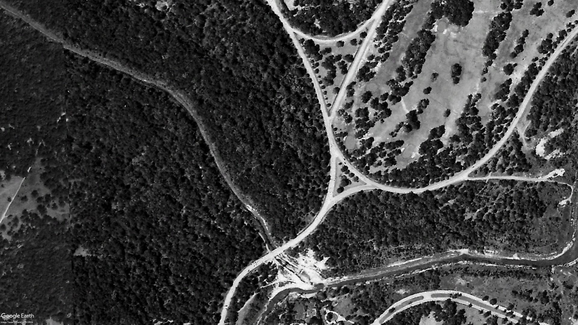 Black-and-white aerial view of a highway interchange cutting through forested terrain; looping ramps form a cloverleaf, with dense trees on one side and lighter open fields on the other.