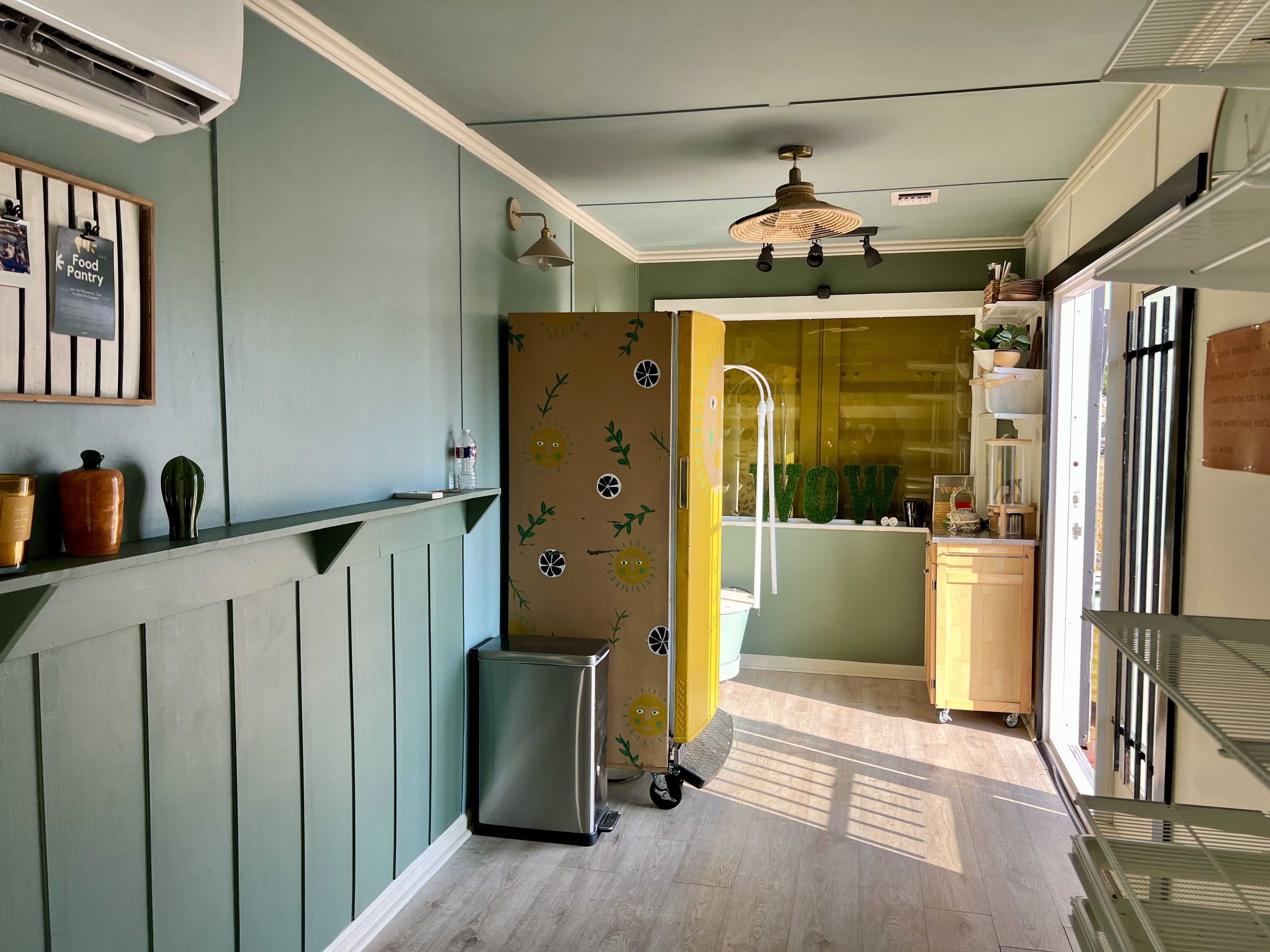 Room with green walls and light wood floor, a yellow painted fridge with sun and leaf designs, small wooden cabinet, metal trash bin, and shelves with decorative pots and plants.
