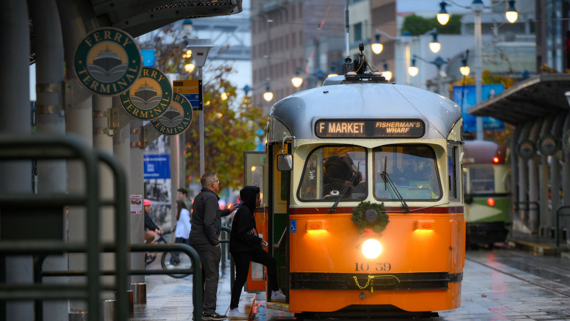 Front of a vintage orange streetcar with display "F MARKET" and "Fisherman's Wharf"; wet pavement, pedestrians boarding, and round ferry-terminal signs on the left.