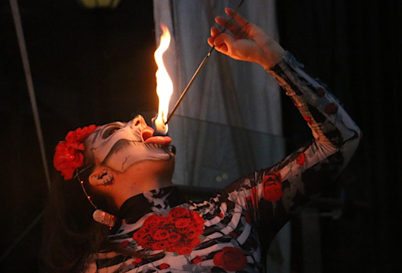 Performer with skeletal face paint and a red flower headpiece breathes fire on stage, holding a flame wand near their open mouth; wearing a floral dress adorned with red roses.