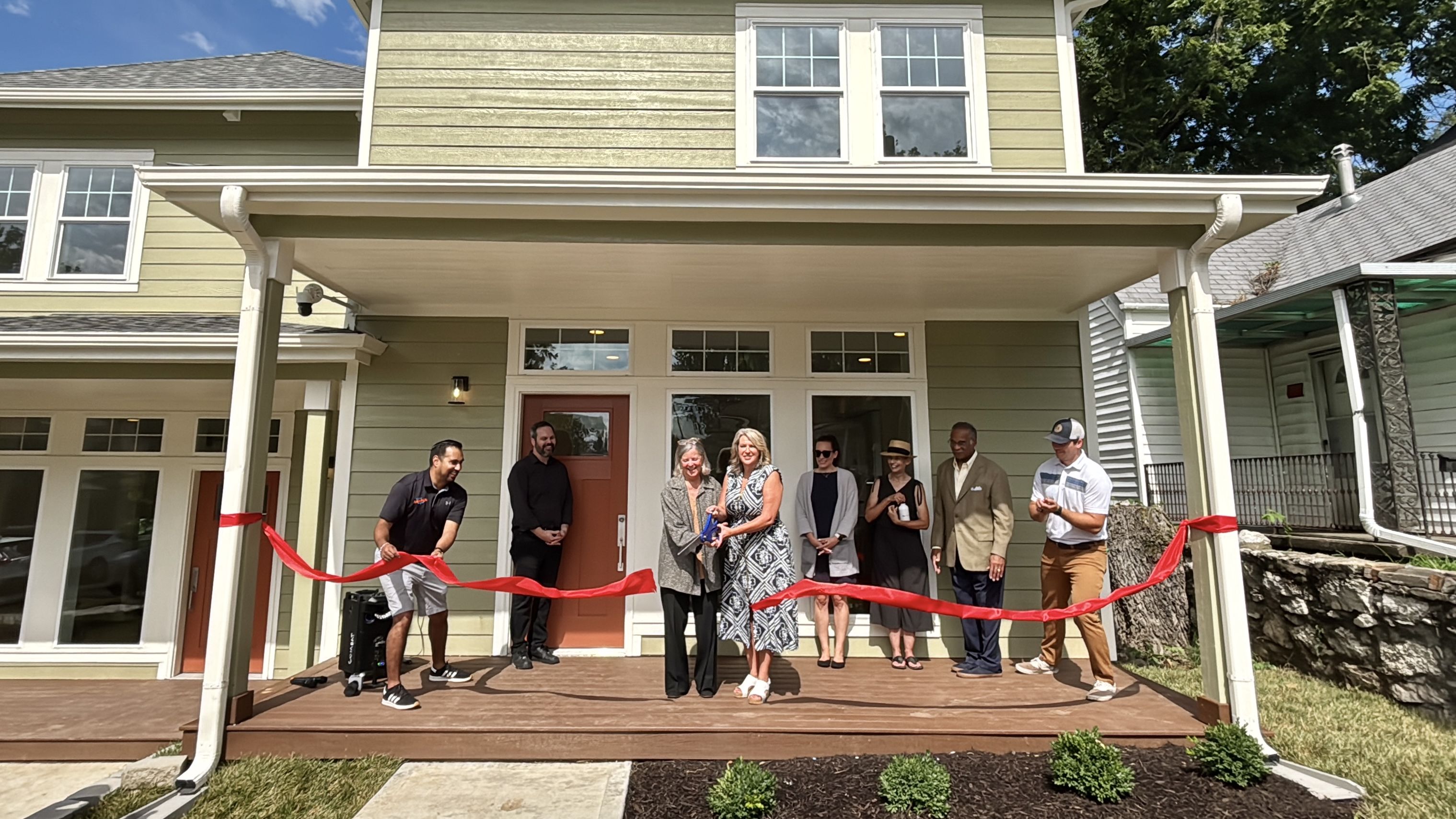 Group of people in smart casual attire stand on a wooden porch in front of a light-green, two-story house with white trim. A red ribbon spans the entry for a ribbon-cutting ceremony.