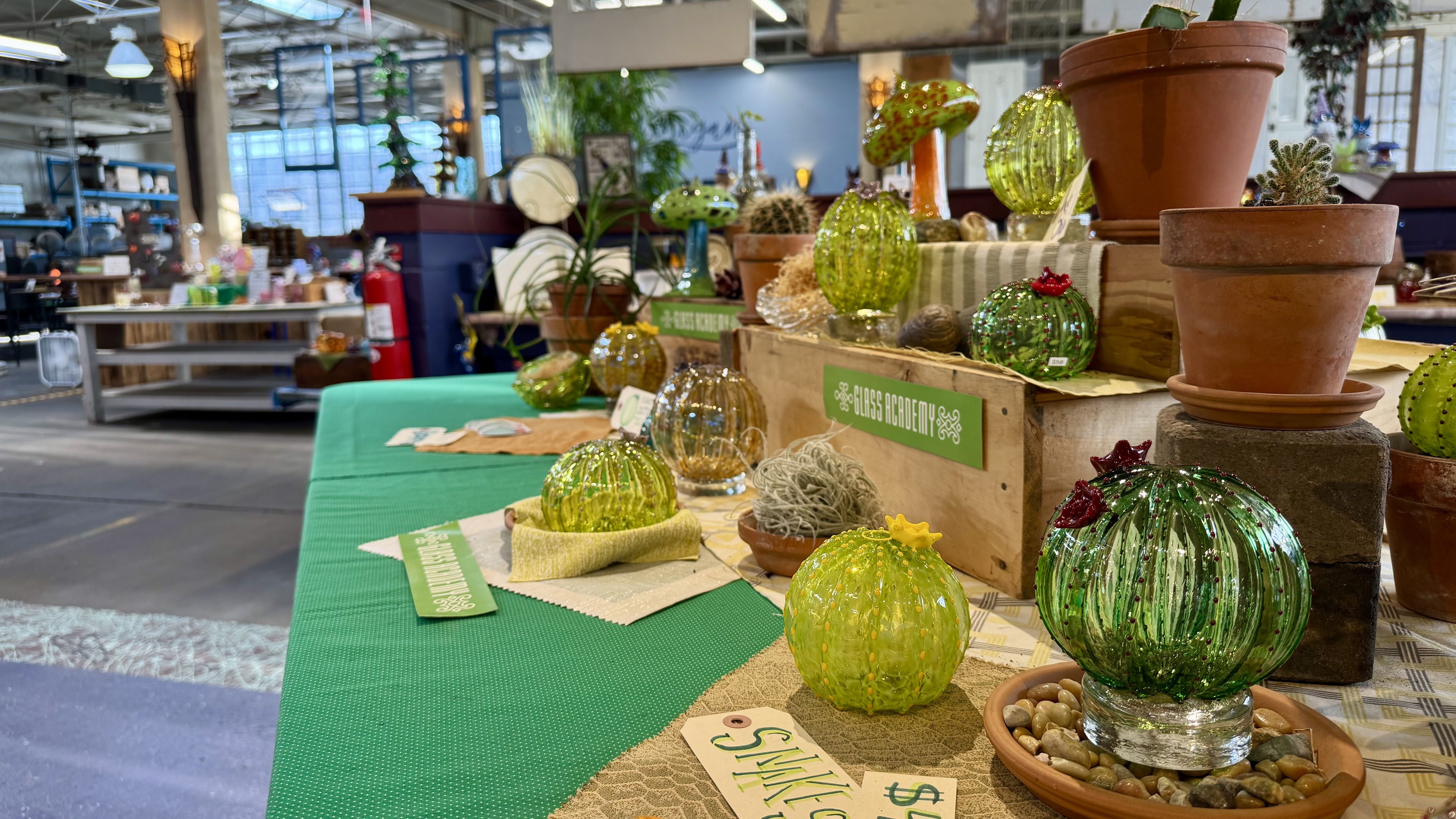 Indoor store in an industrial-looking building with glass cacti and green glass orbs on a display table, surrounded by terracotta pots and potted plants; shelves and a "Glass Academy" sign in the background.