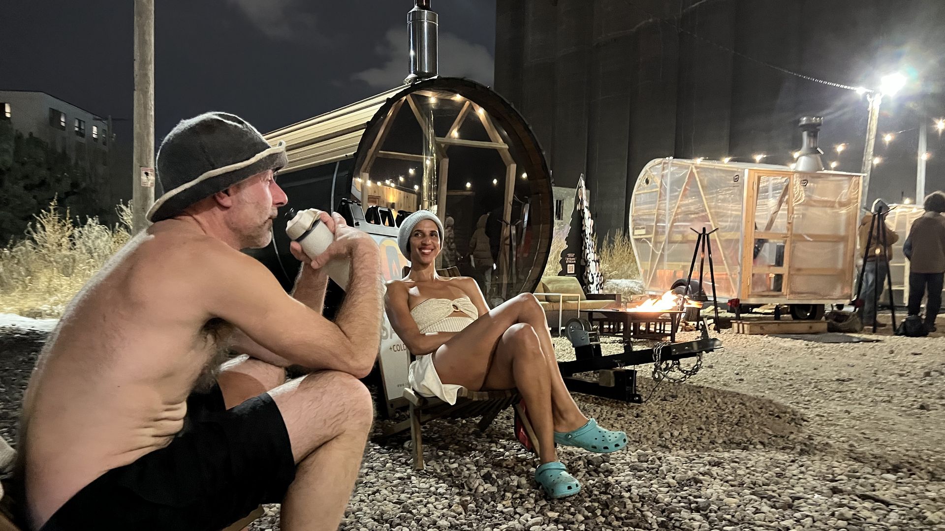 A man and a woman sitting outdoors in bathing suits near two saunas. One sauna is barrel-shaped and black, the other is made of translucent material with a wood frame.
