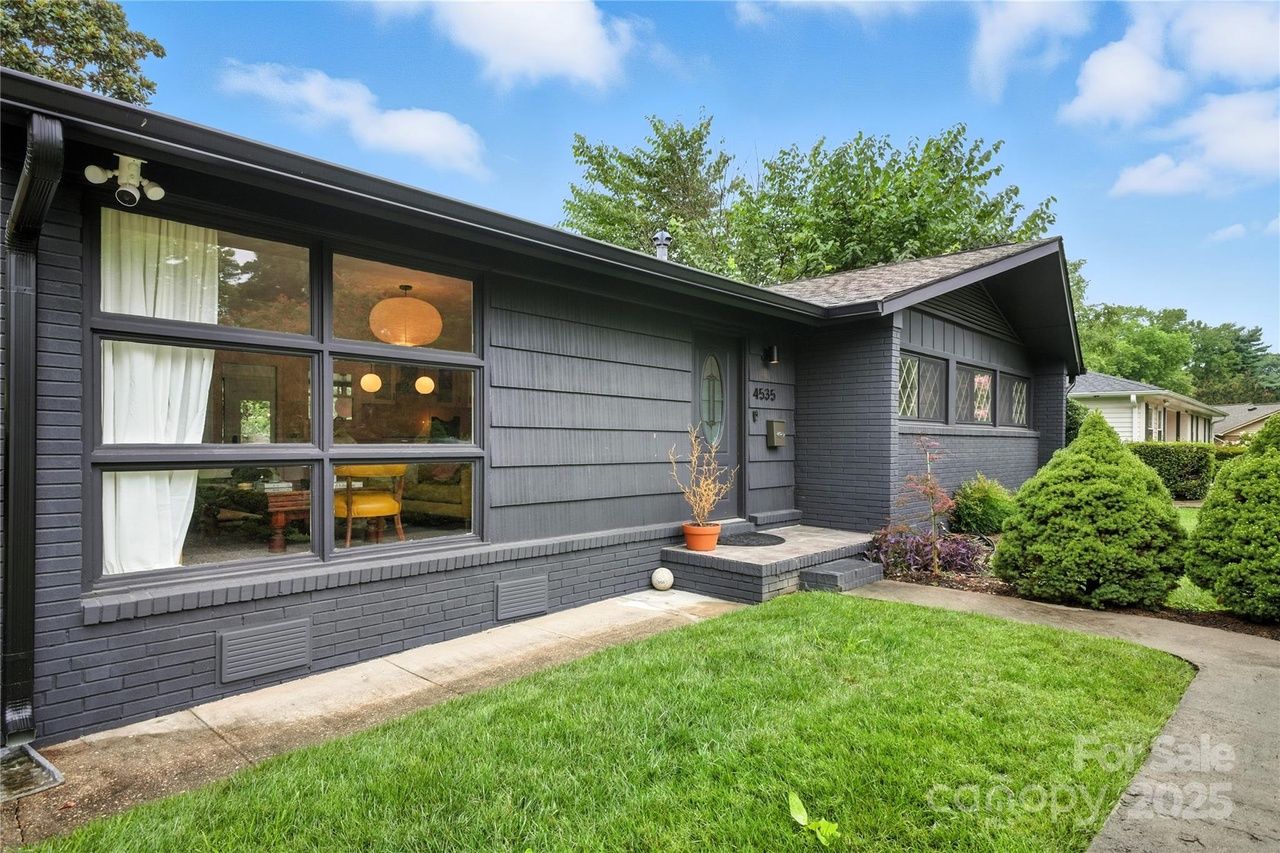 Dark gray single-story house with large windows, green lawn, small shrubs, and a curved concrete walkway under a blue sky with scattered clouds.